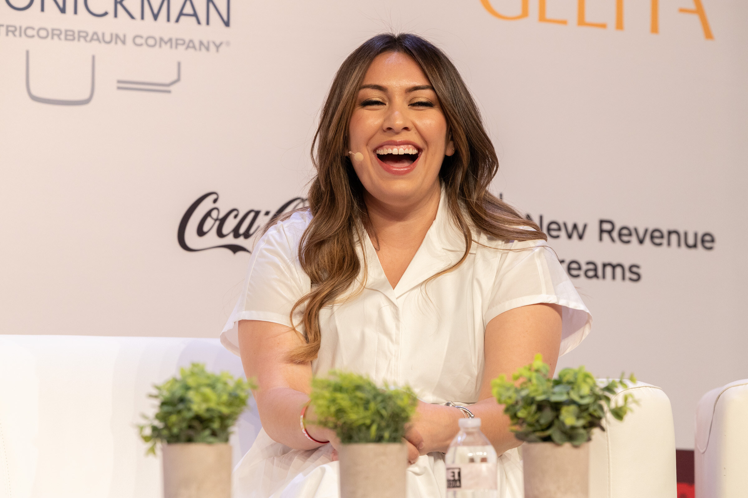 Laughing woman with long brown hair in a white shirt seated behind small potted plants on stage.
