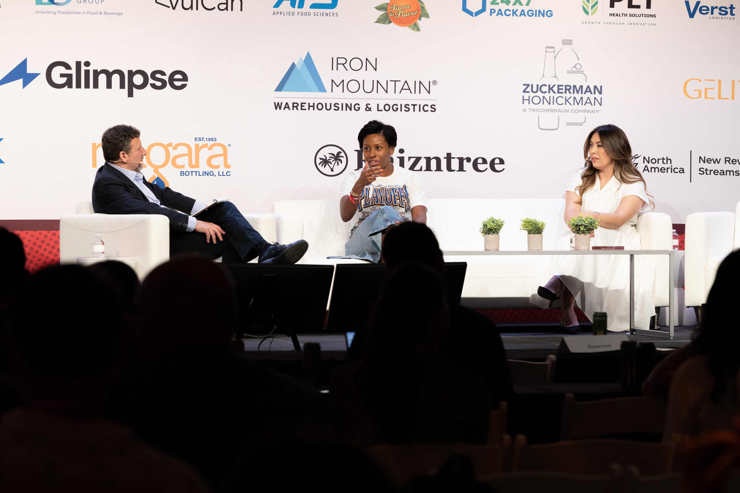 Three people seated on white couches on stage in front of a sponsor wall, with audience silhouettes in front.