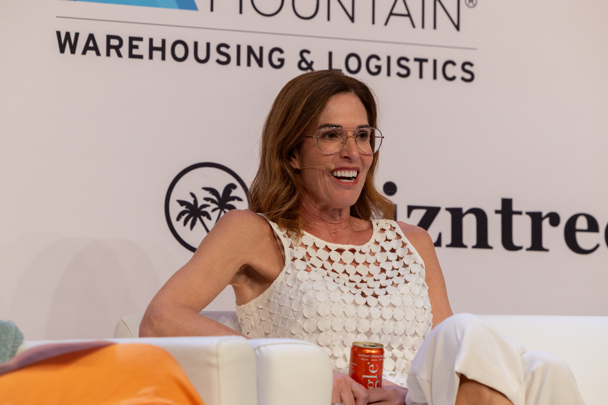 Smiling woman in glasses and a white crochet top seated on a couch holding an orange can.