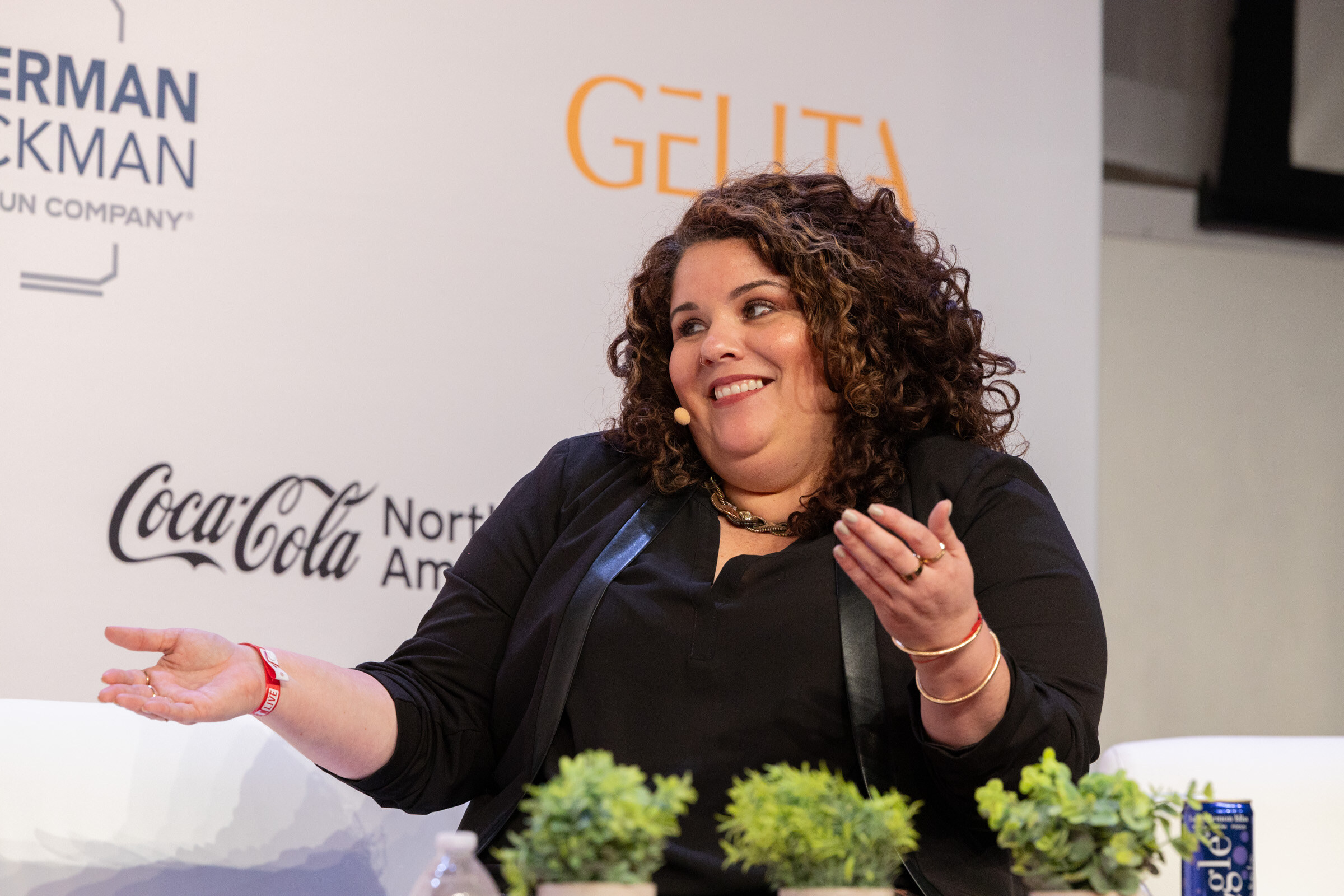 Smiling woman with curly hair in a black jacket gesturing with both hands, in front of a Coca-Cola North America logo.