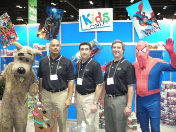Three men in black polo shirts stand between a costumed dog character and a person in a Spider-Man costume. The Kids Only sign and superhero artwork hang behind them.