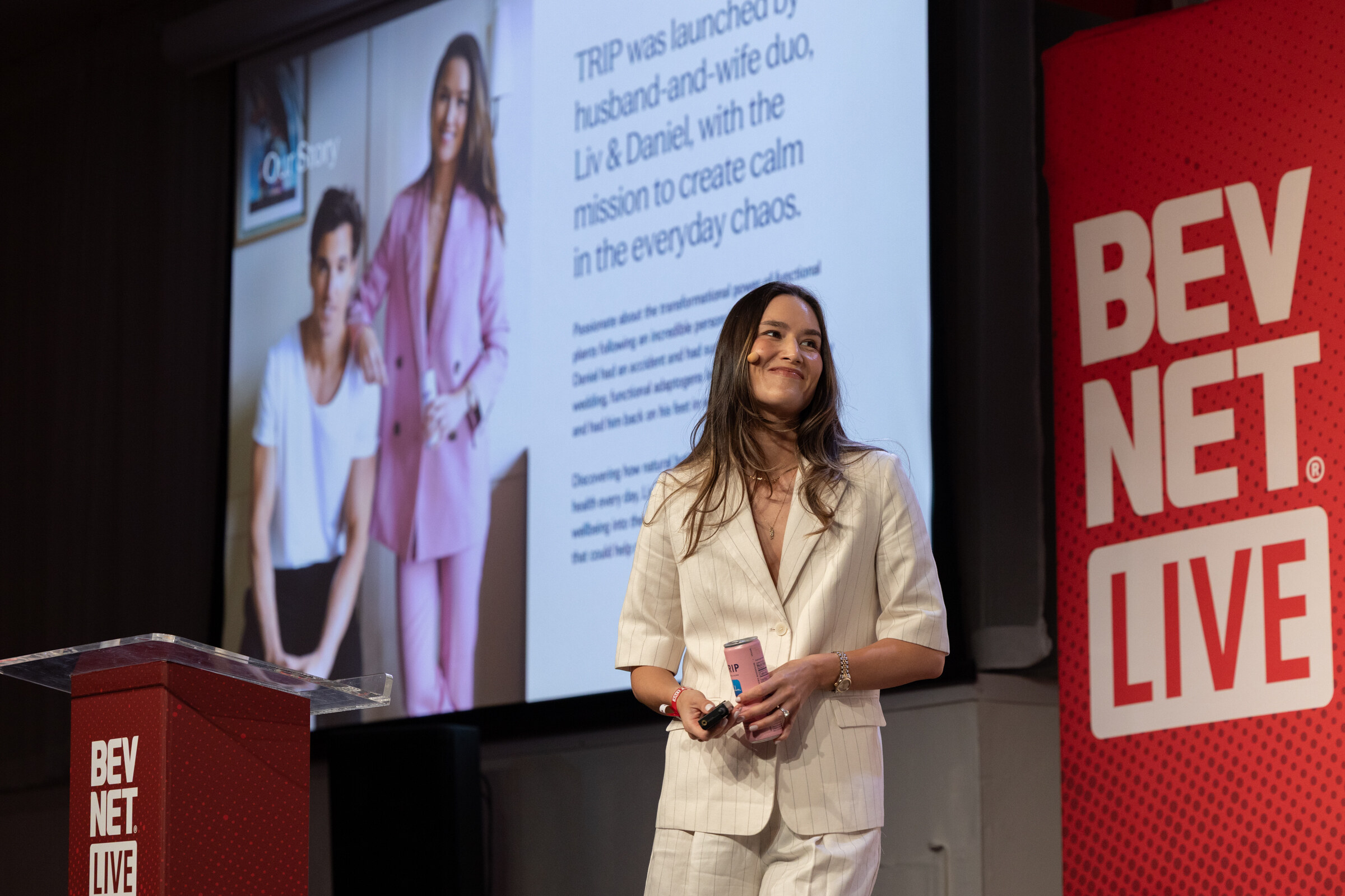 Woman in a cream pinstripe suit holding a pink TRIP can on stage beside a slide about the brand.