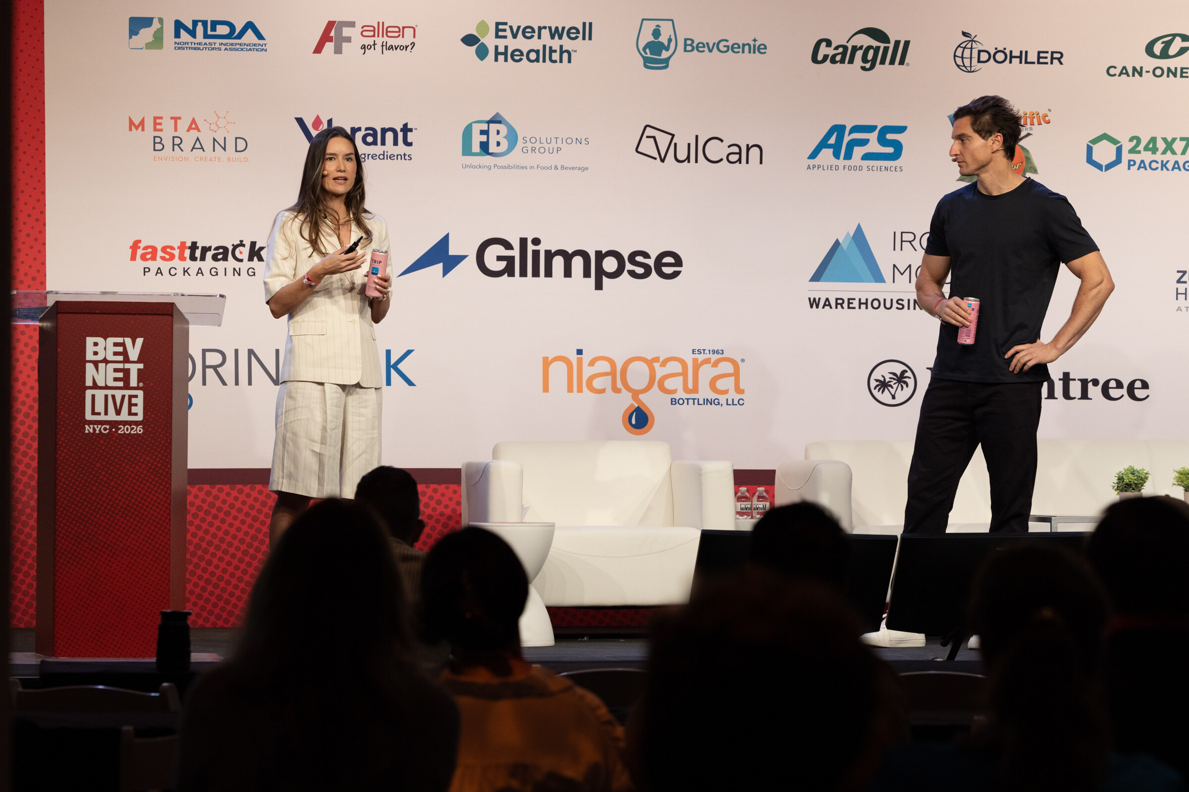 Woman in a cream suit speaking and a man in a black T-shirt holding a pink can, standing in front of a sponsor wall.