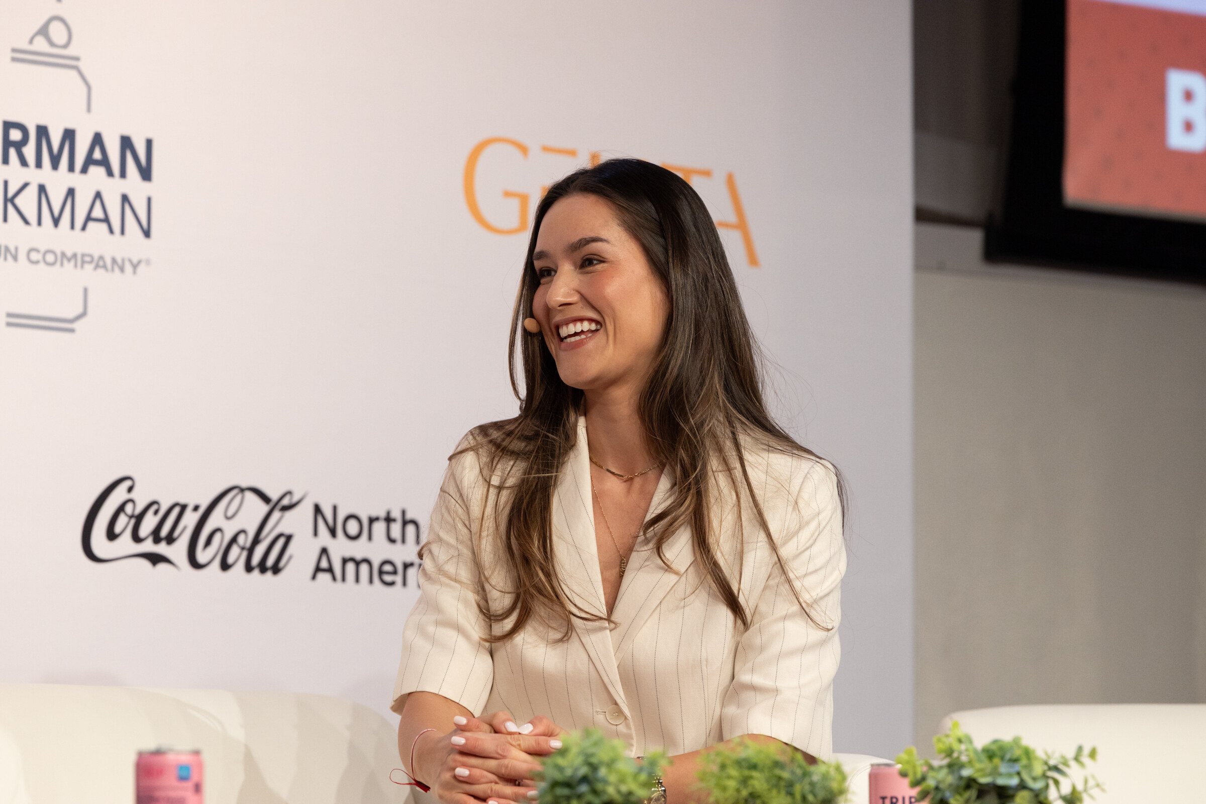 Smiling woman with long dark hair in a cream blazer seated on stage, a pink can on the table in front of her.