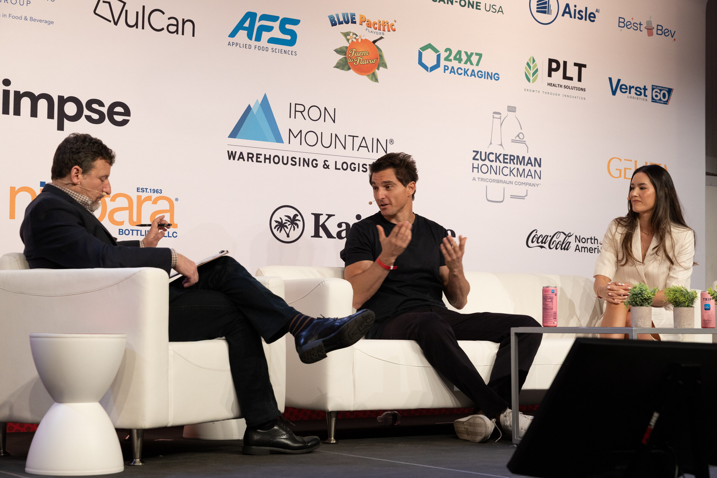 Man with notes, a man in a black T-shirt gesturing and a woman in a cream blazer seated on white couches.