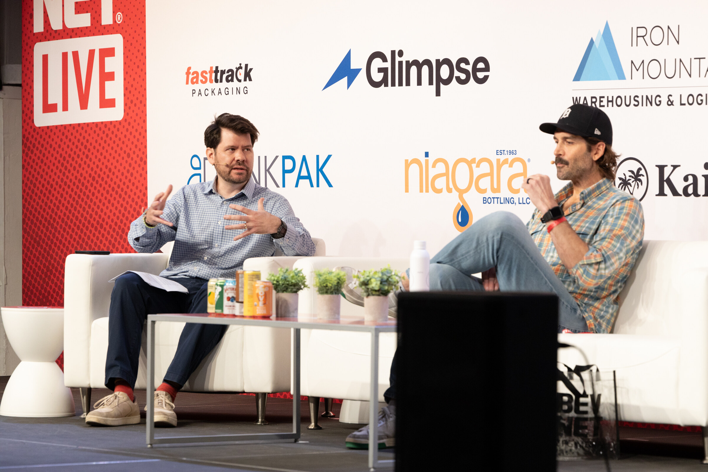 Man in a checked shirt gesturing toward a man in a cap and plaid shirt, with cans on a low table in front of a Niagara logo.