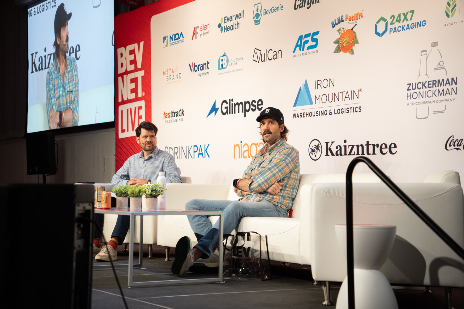 Two men seated on white couches on stage, one in a cap and plaid shirt, with a large screen showing the speaker above.