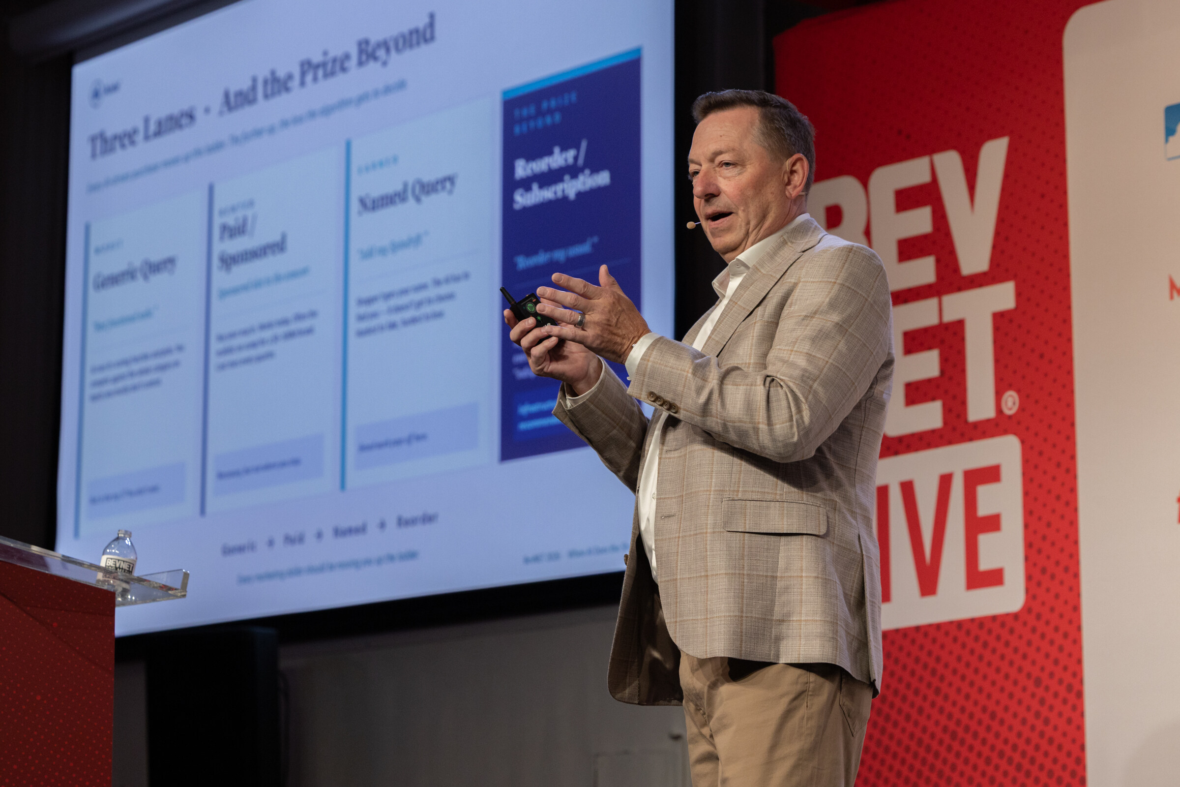 Man in a tan plaid blazer gesturing with a clicker beside a projected slide titled Three Lanes and the Prize Beyond.