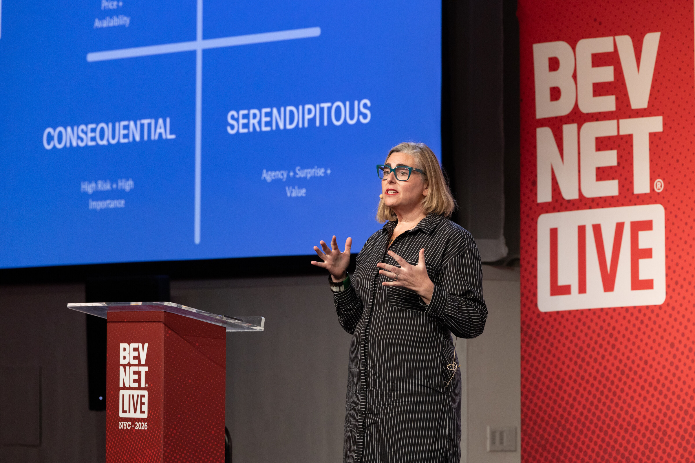 Woman in glasses and a striped shirt dress speaking beside a podium in front of a blue slide reading Consequential and Serendipitous.