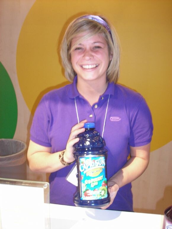 A smiling woman with blonde hair and a purple headband, wearing a purple Kraft polo, holds a large bottle of Capri Sun juice at a booth with a yellow backdrop.