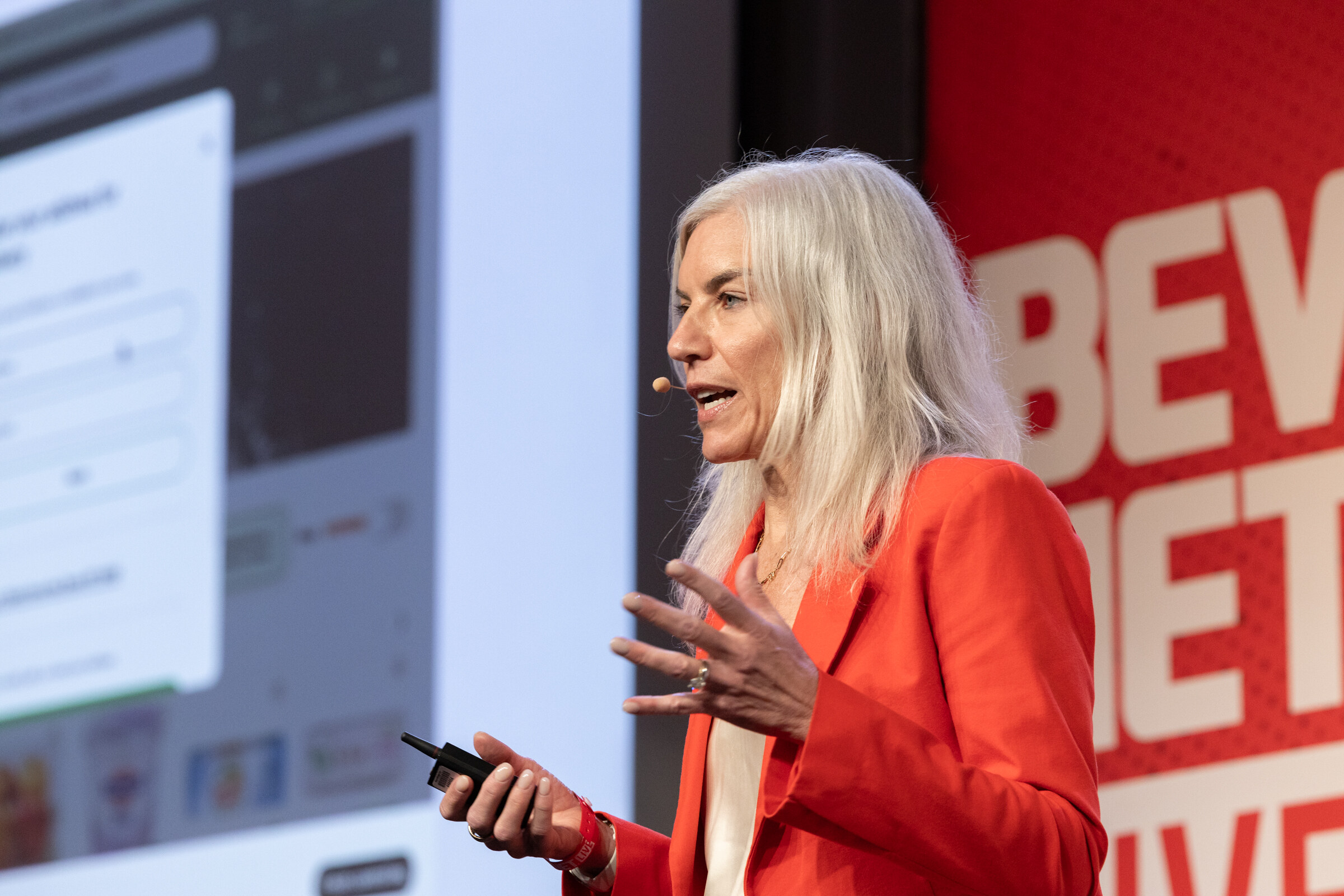 Woman with long silver hair in a red blazer speaking and gesturing, a clicker in hand.