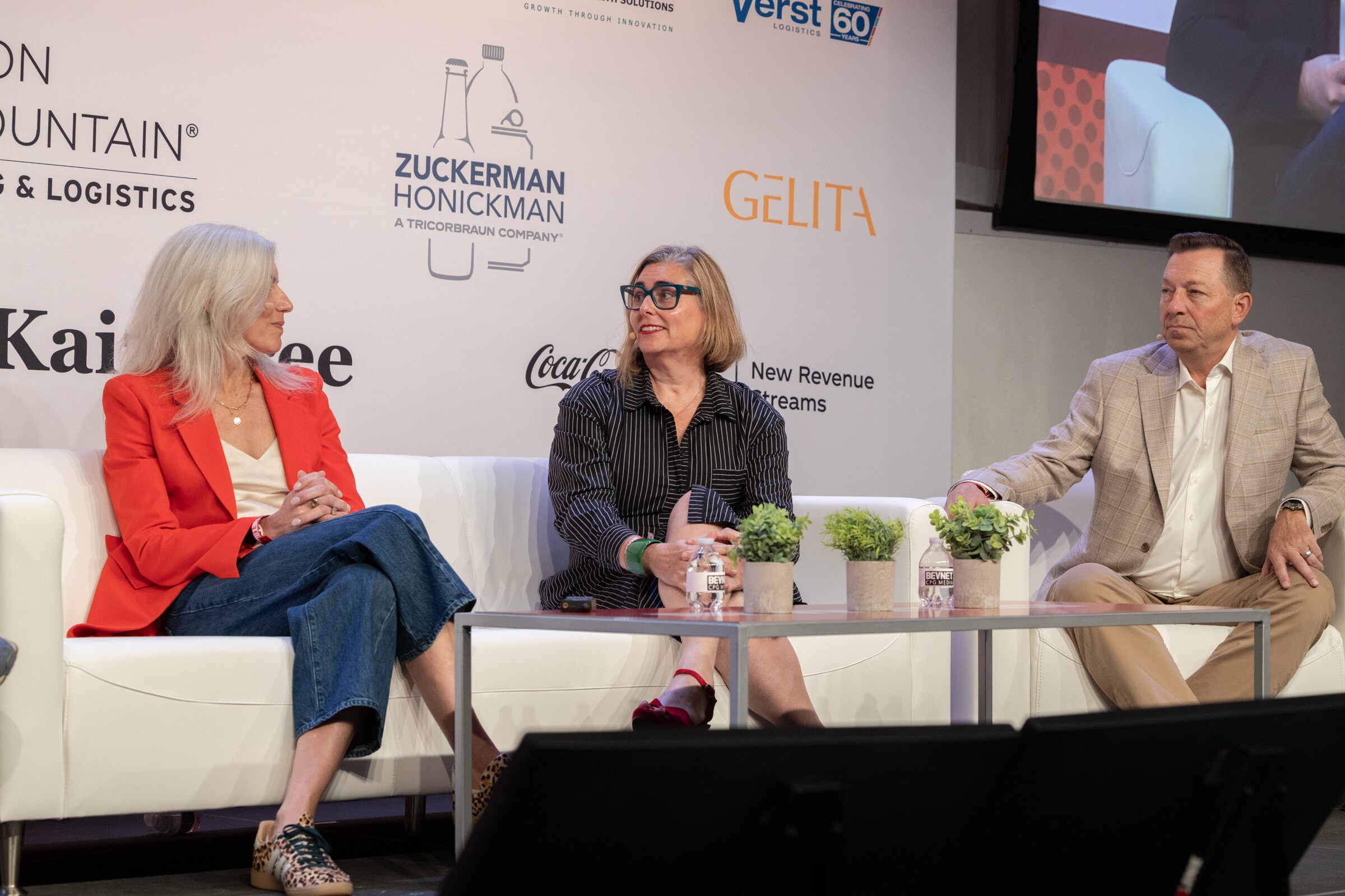 Two women and a man seated on white couches on stage in front of a sponsor wall.