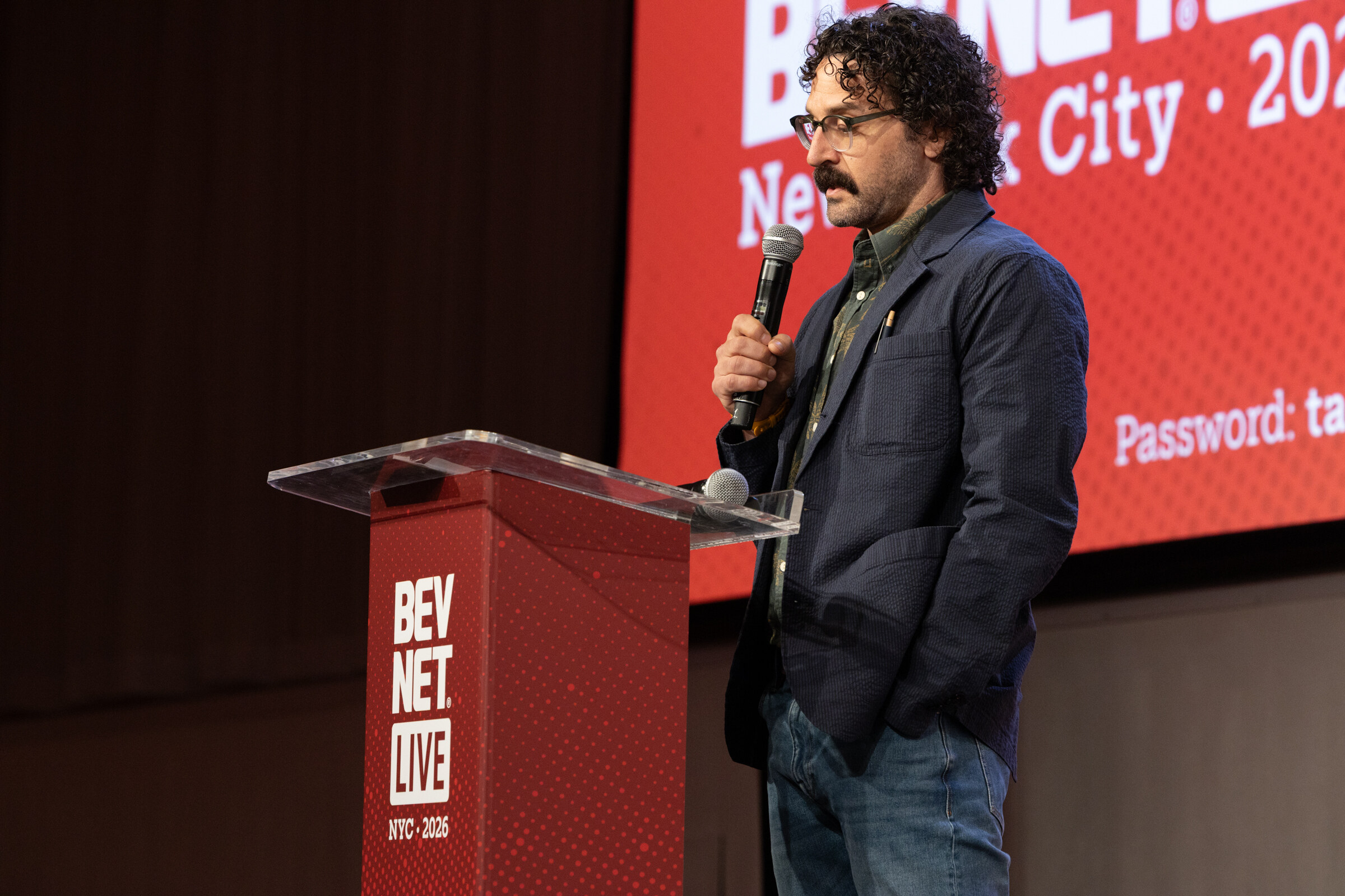 Man with curly hair and glasses holding a microphone beside a red BevNET Live podium.