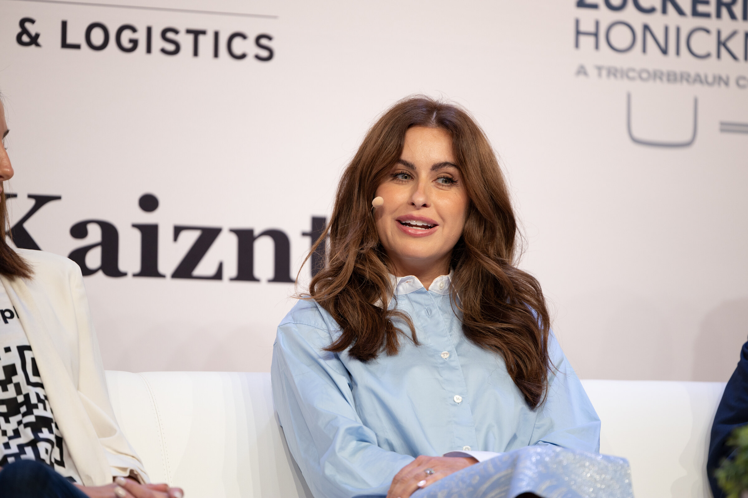 Woman with long wavy brown hair in a light blue shirt seated on a white couch on stage.