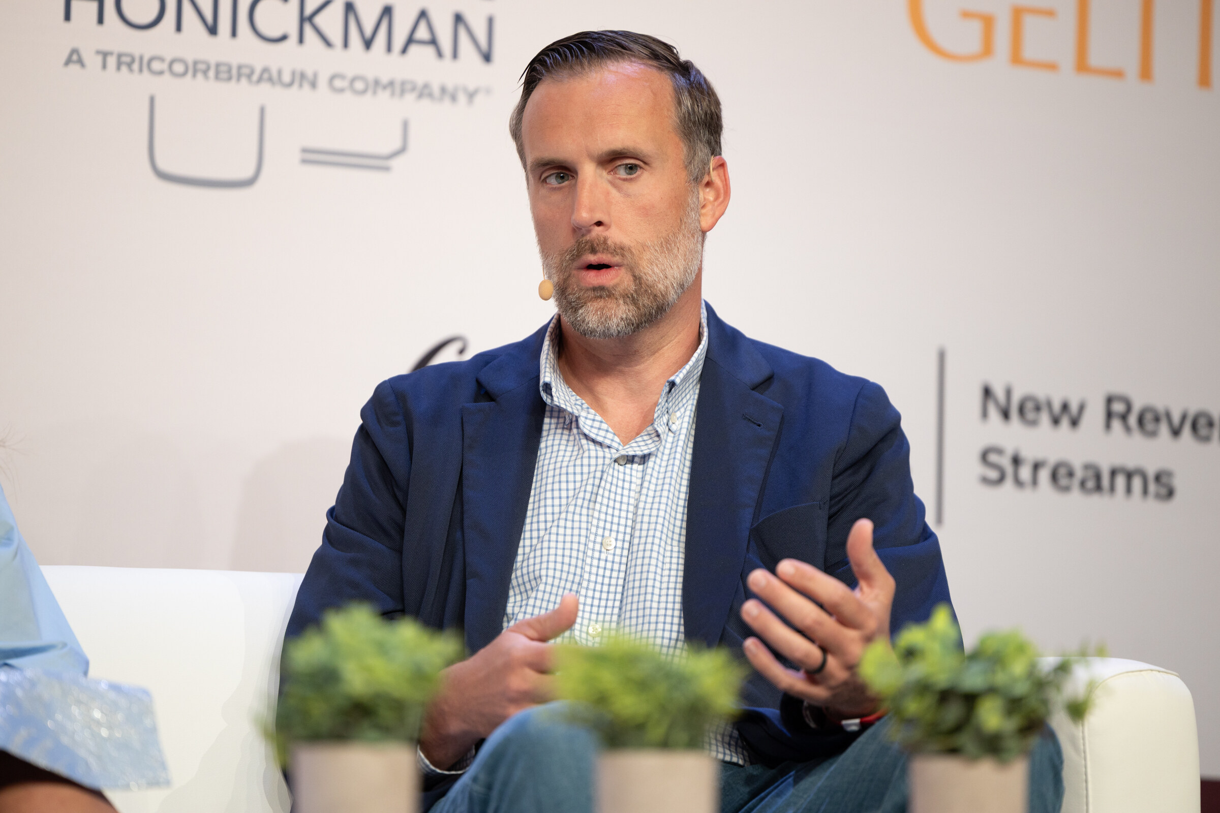 Man with a beard in a navy blazer speaking and gesturing behind small potted plants.