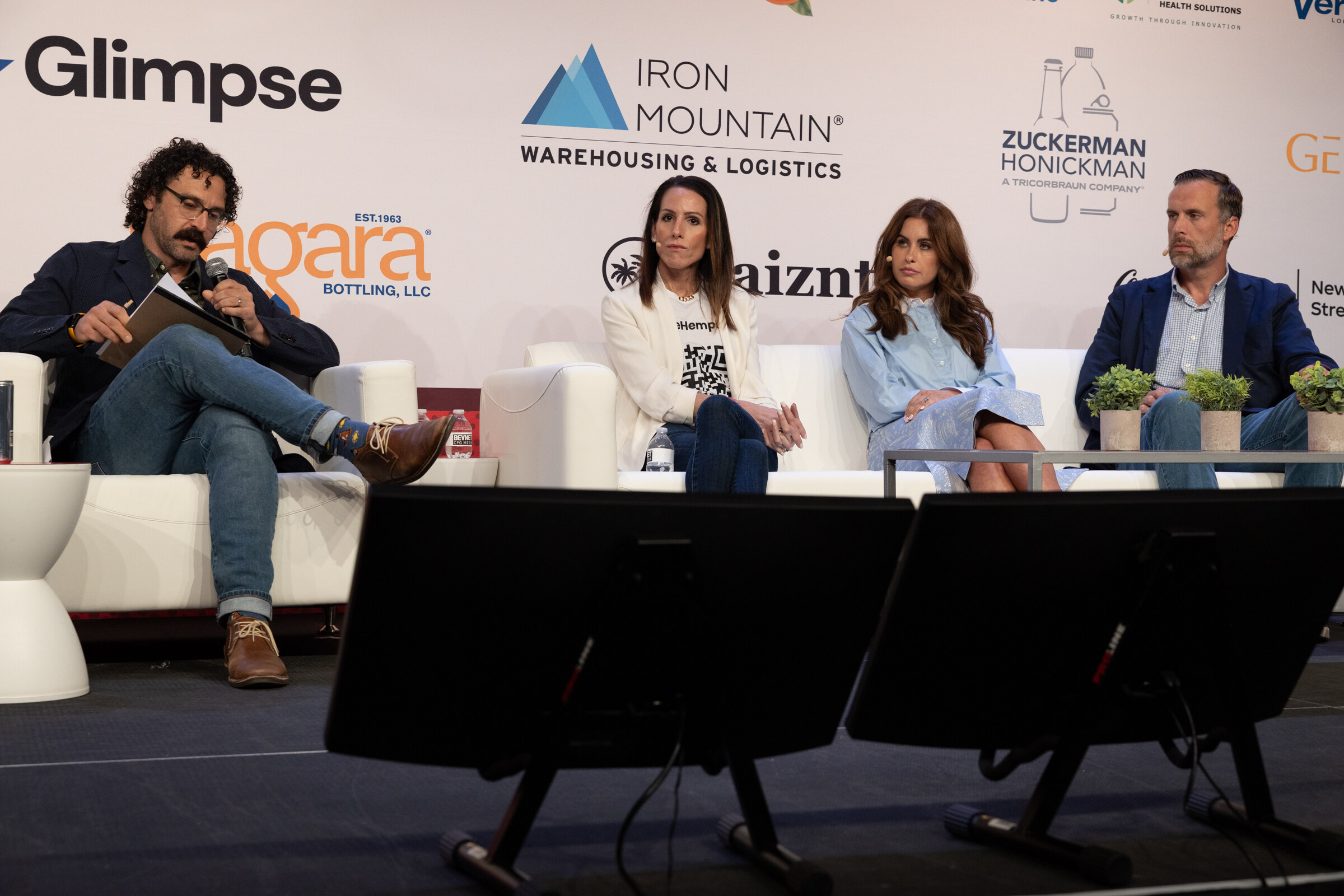 A moderator with notes and three panelists seated on white couches on stage in front of a sponsor wall.