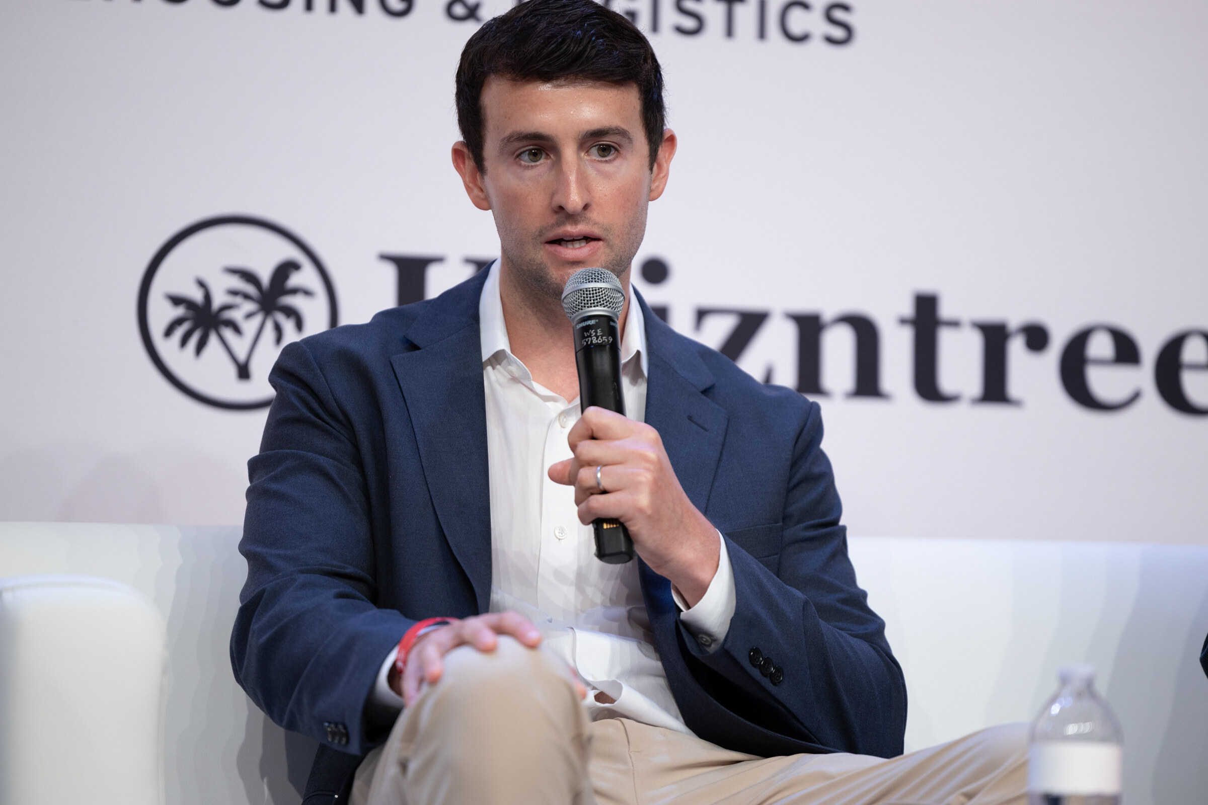 Man in a navy blazer and white shirt speaking into a handheld microphone on stage.