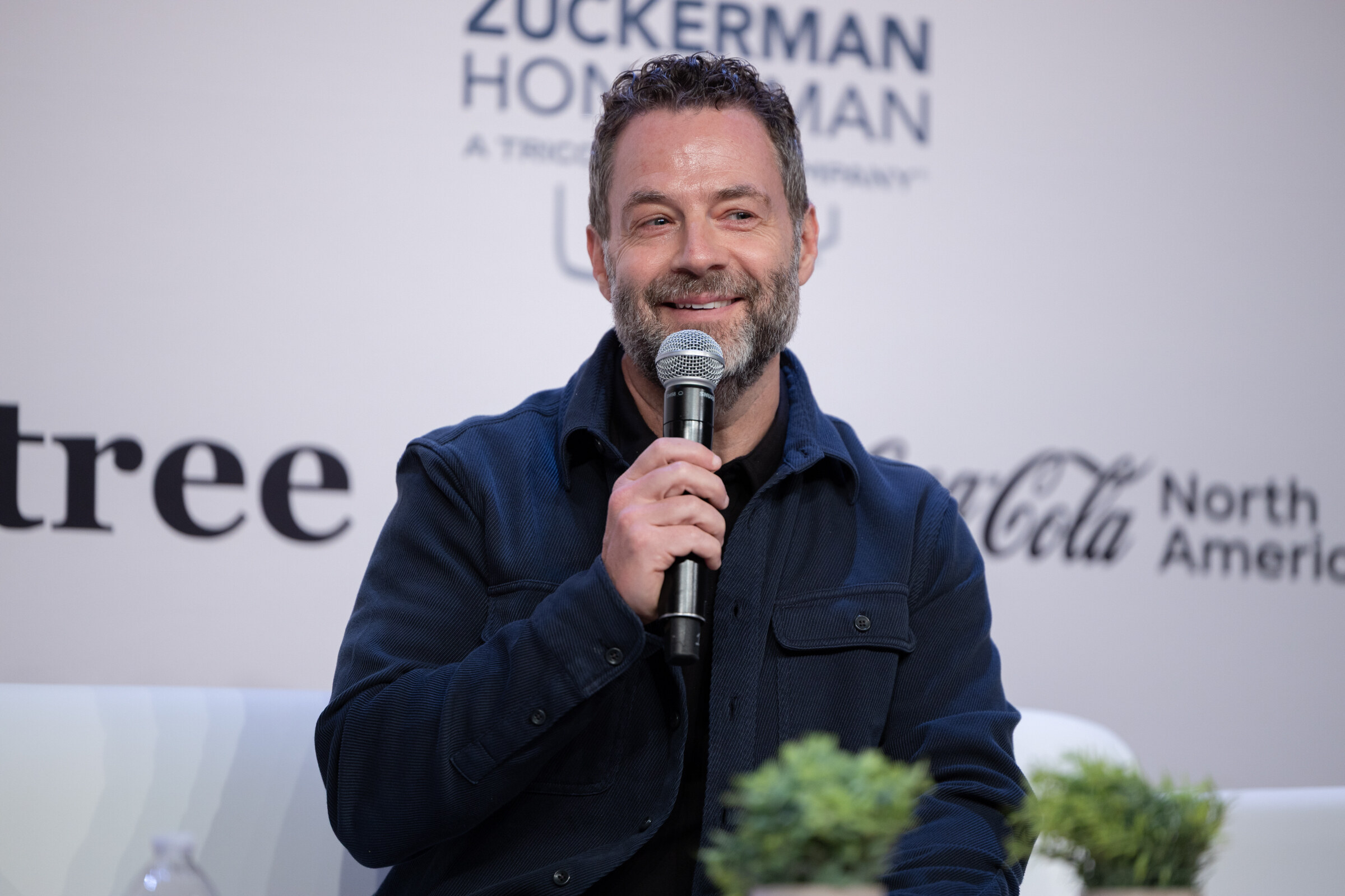 Smiling man with a beard in a dark jacket holding a microphone in front of a Coca-Cola North America logo.