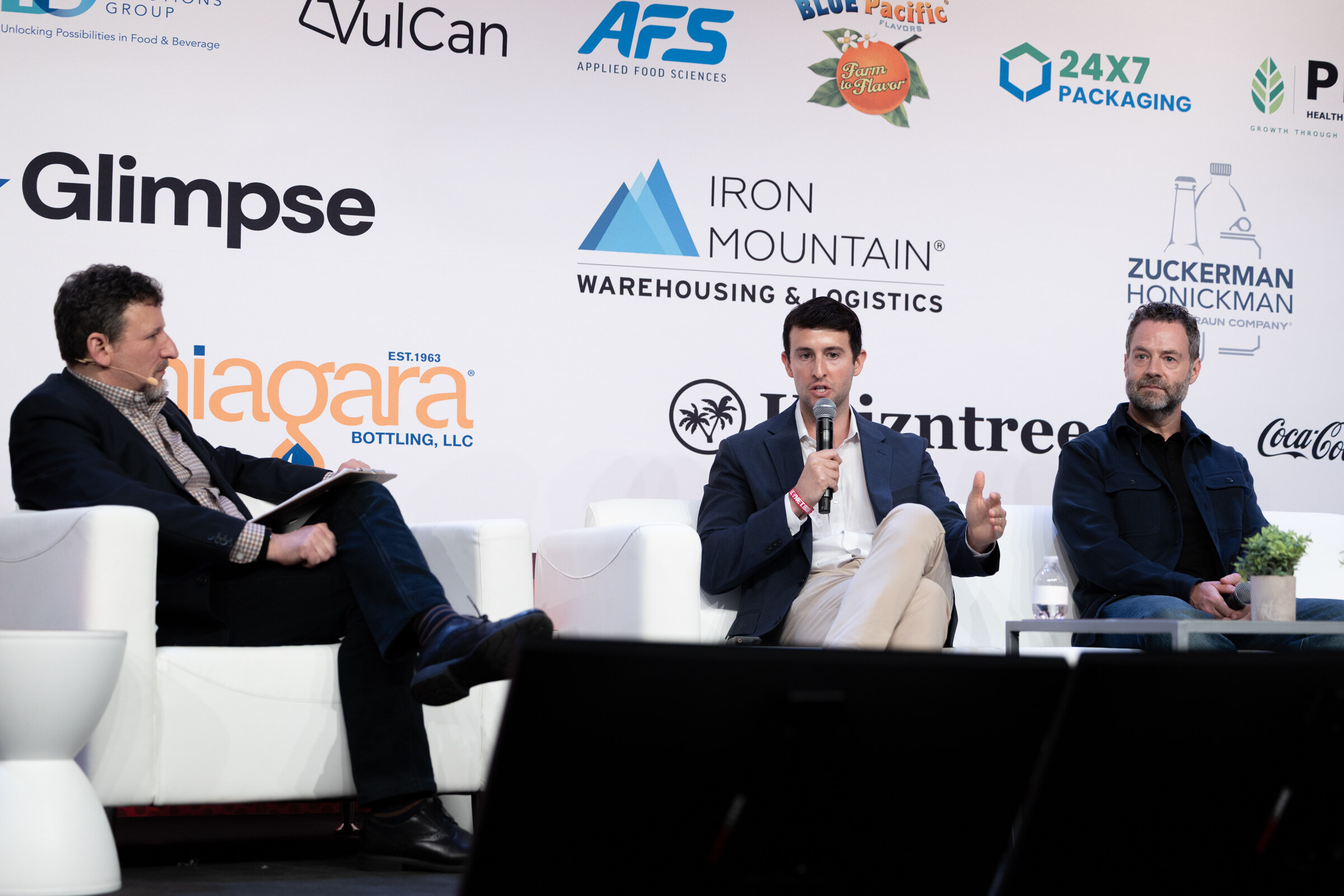 A moderator and two panelists seated on white couches in front of a sponsor wall.