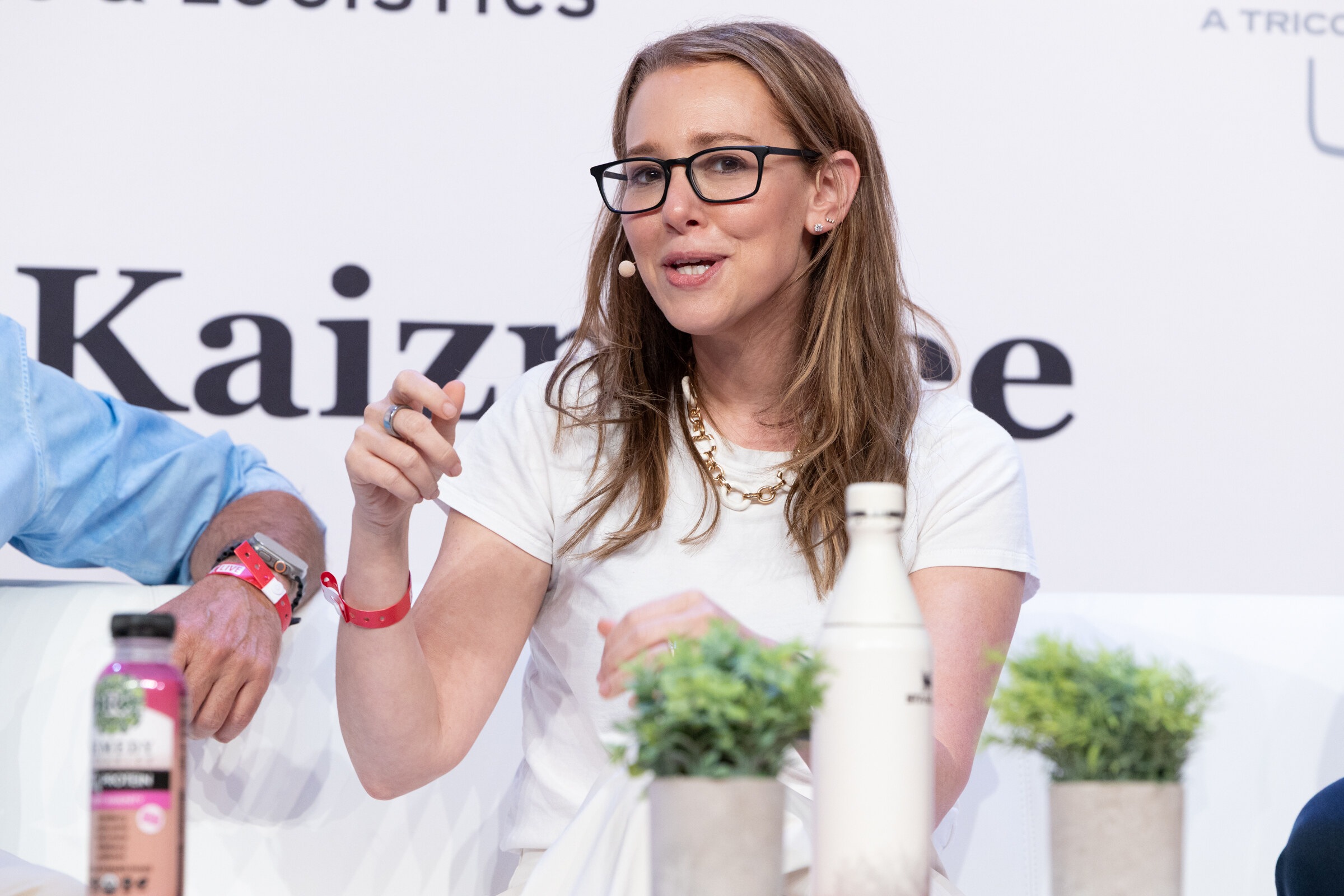 Woman in glasses and a white T-shirt speaking and gesturing on stage, a bottle and potted plants beside her.