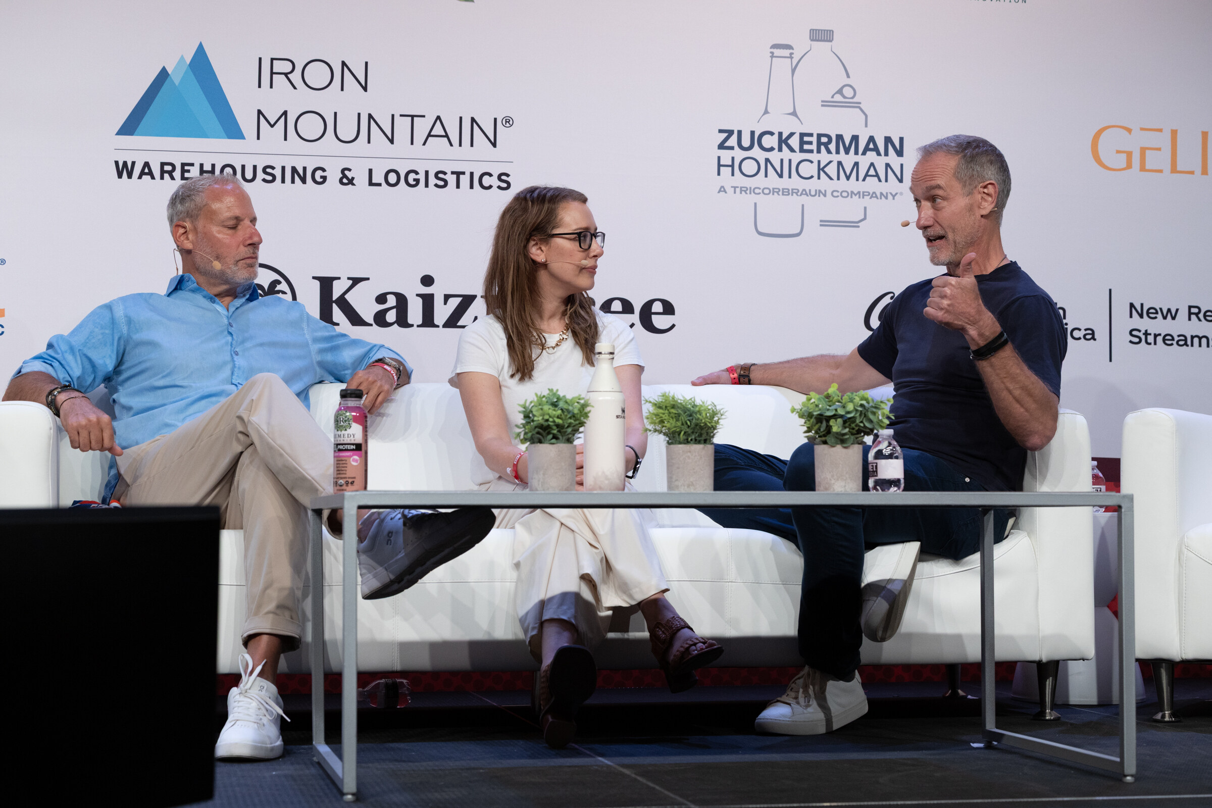 A man in a blue shirt, a woman in white and a man in a navy T-shirt seated on white couches, one gesturing.