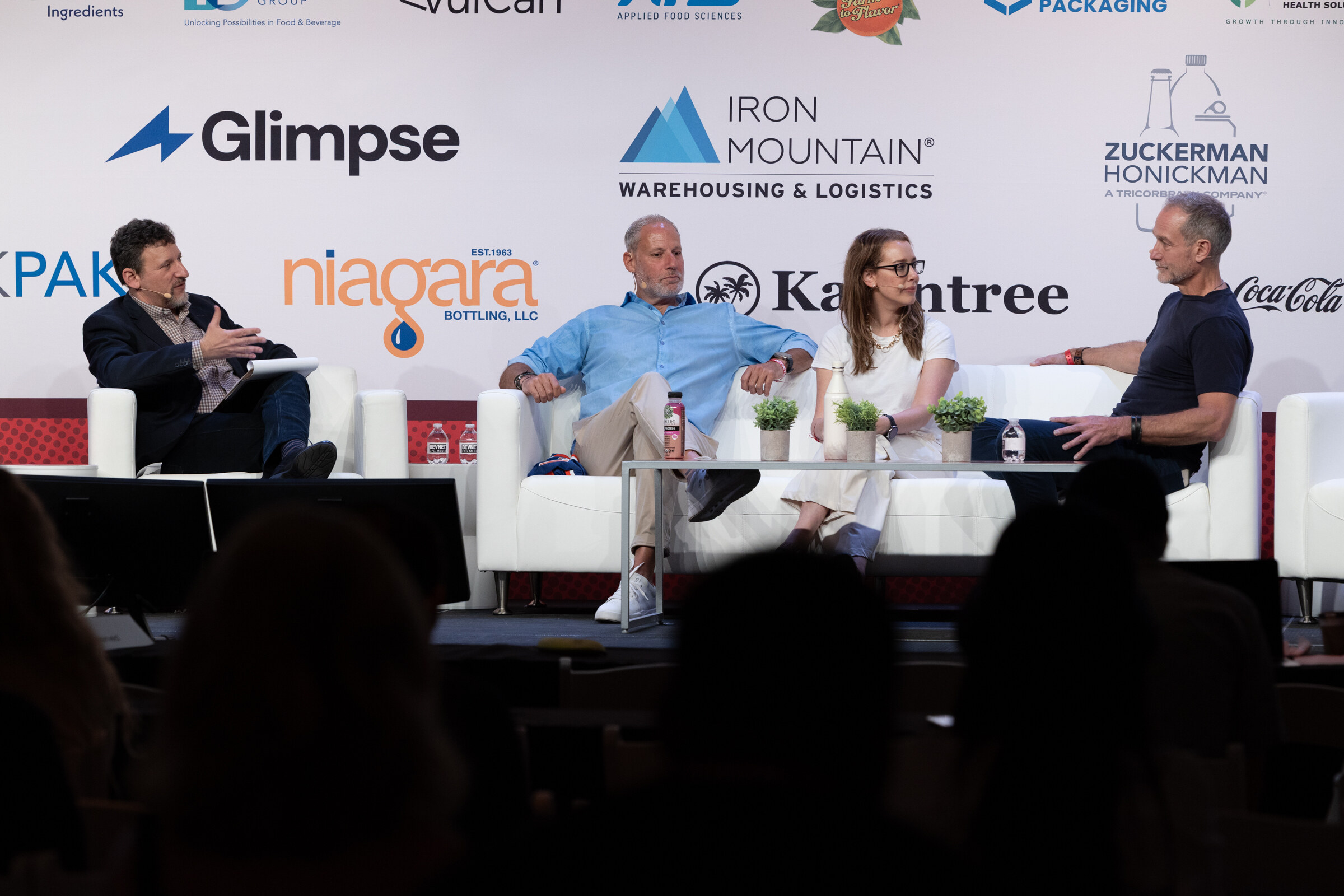 A moderator and three panelists seated on white couches in front of a sponsor wall, with audience silhouettes in the foreground.