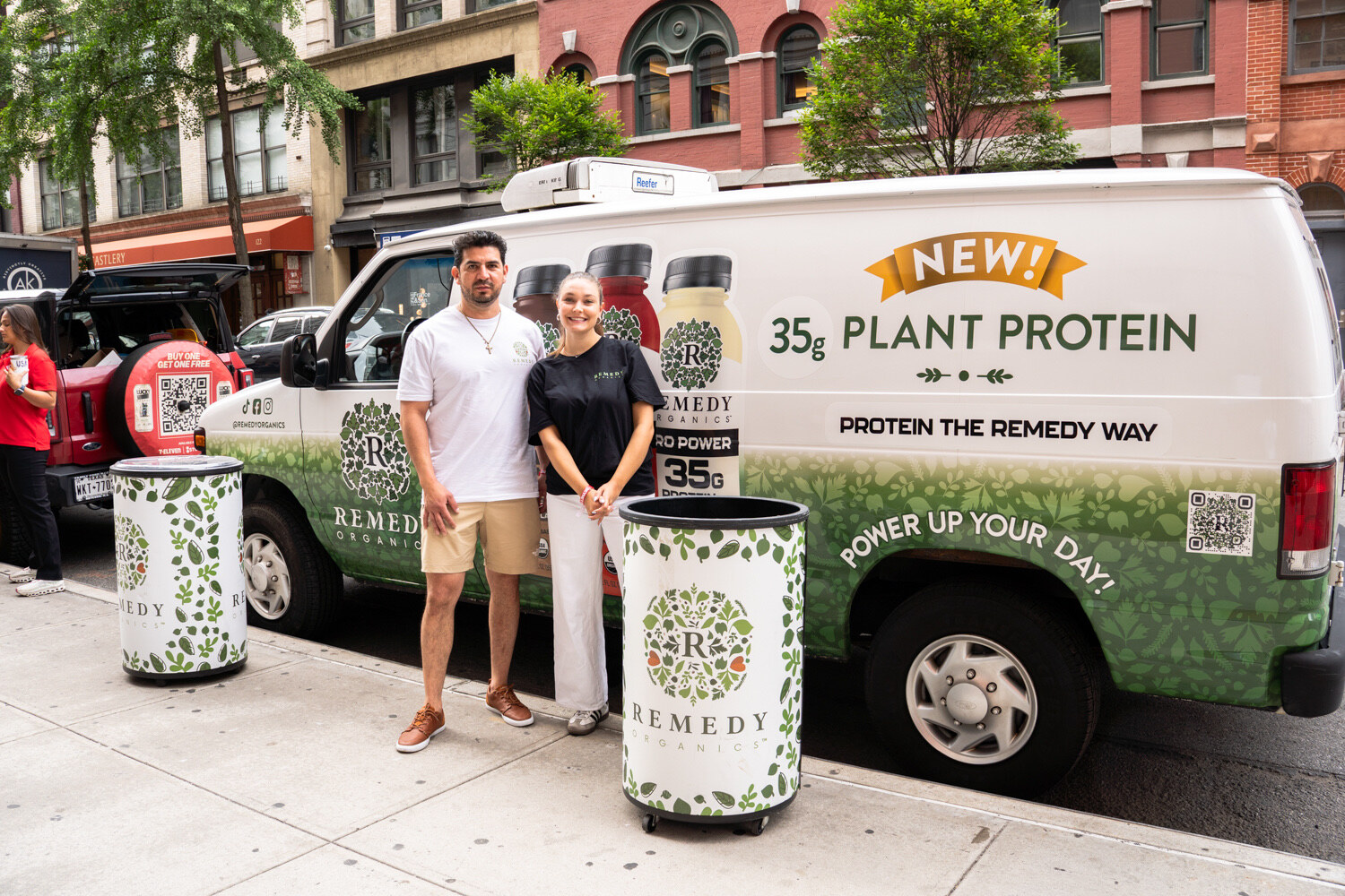 Man and woman stand smiling beside a white van wrapped with Remedy Organics bottles and "New! 35g Plant Protein," next to a leaf-print cooler.