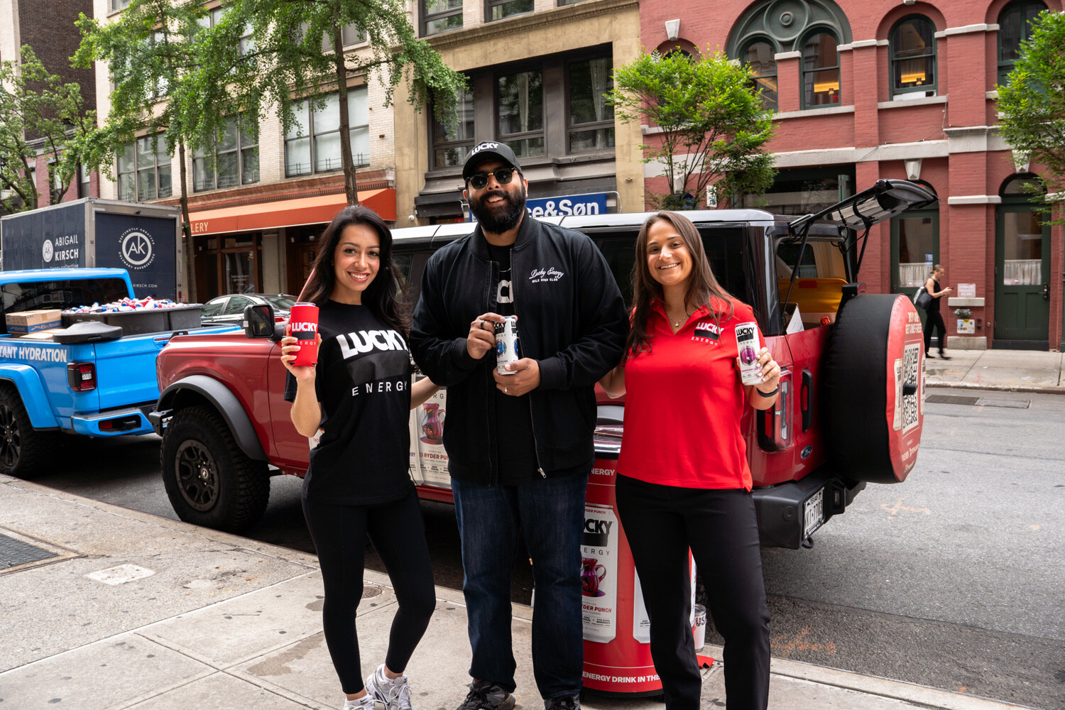 Three smiling people hold Lucky Energy cans next to a red SUV and a red Lucky cooler.