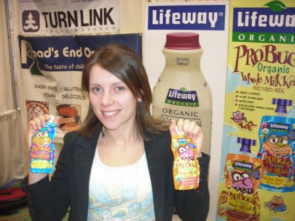 A smiling woman in a dark blazer holds up two colorful ProBugs pouches in front of Lifeway Organic banners and a bottle graphic; a Turn Link banner hangs at the top left.