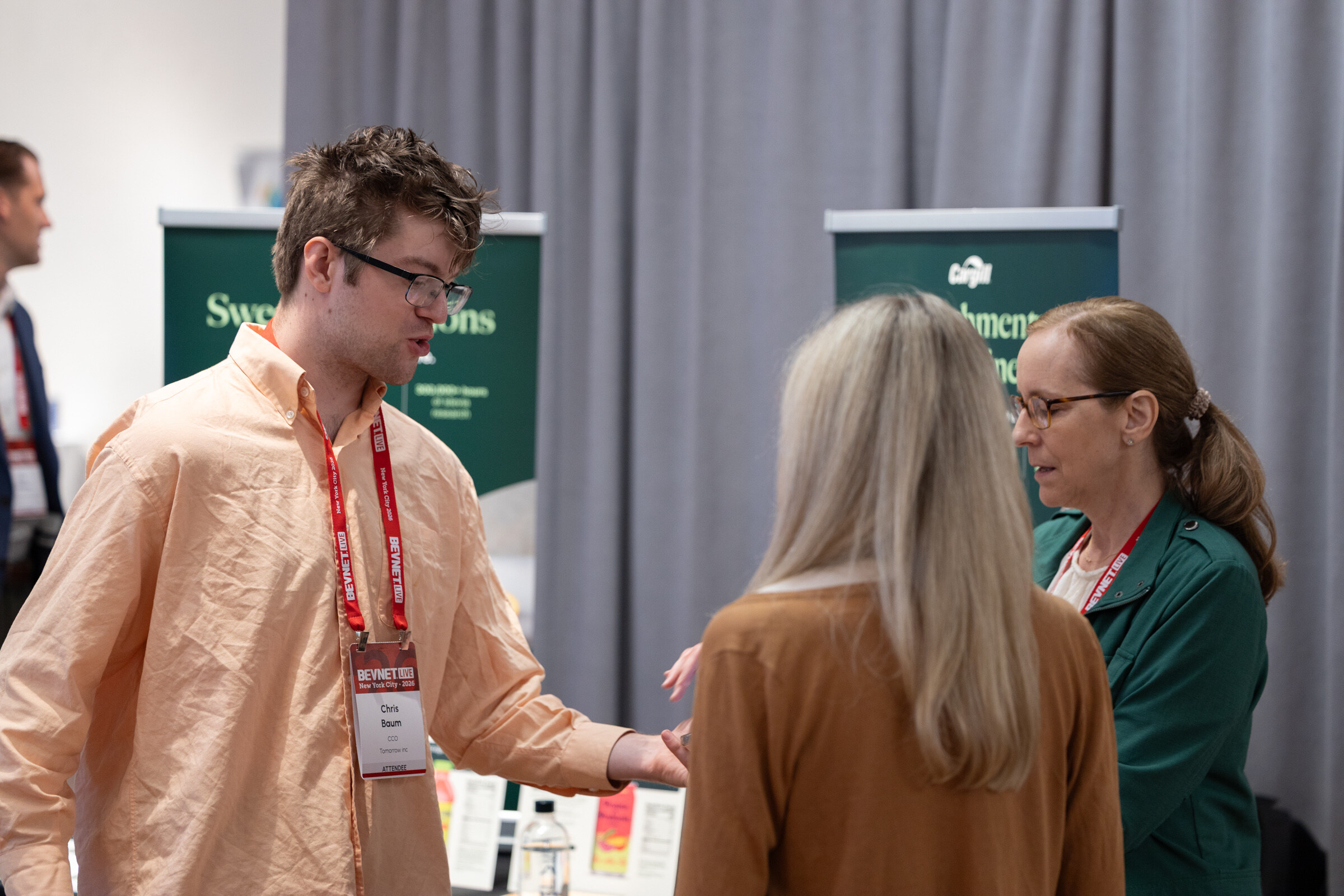 Man in a peach shirt gestures while talking with two women, one in a green jacket, in front of green Cargill banners.