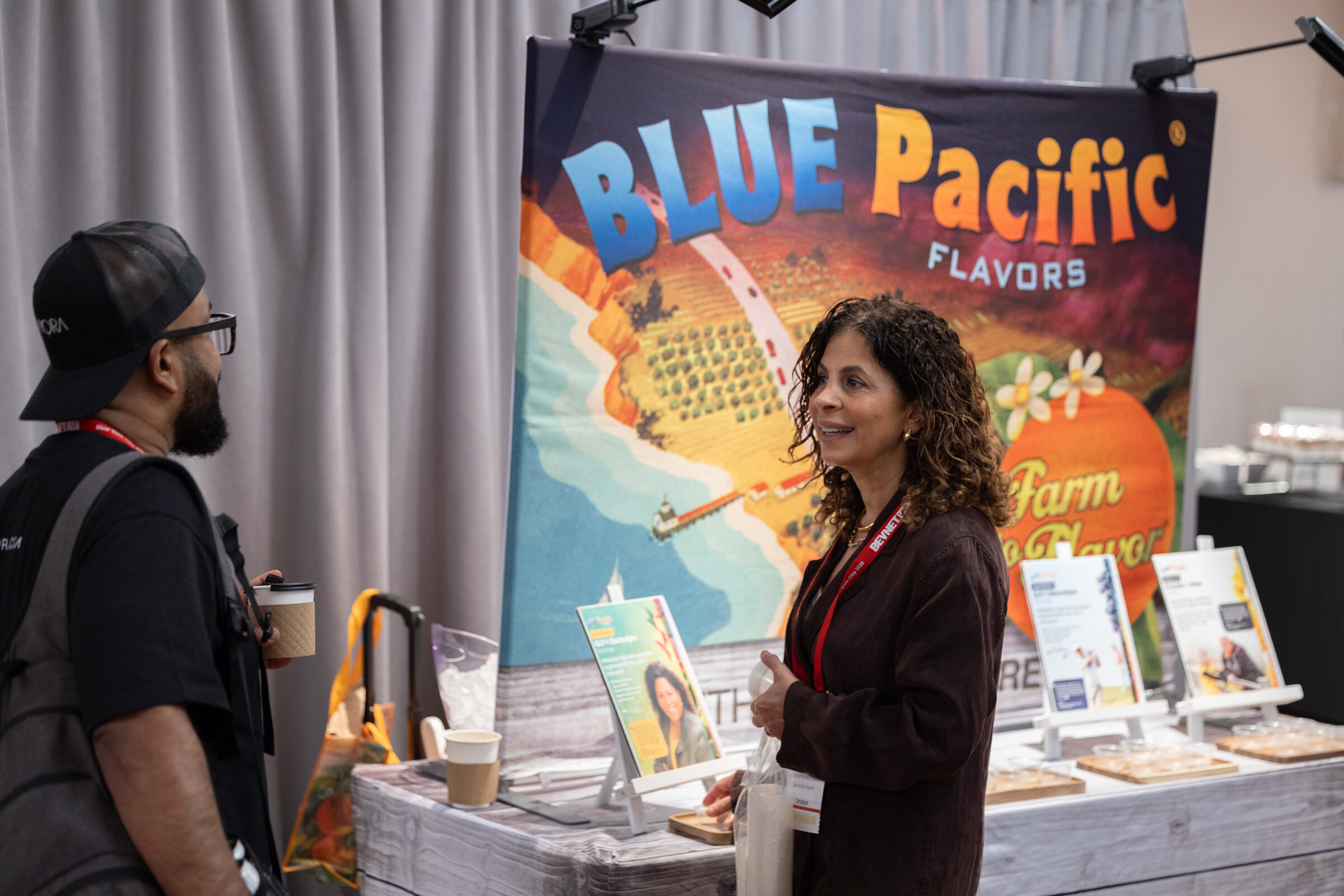 Smiling woman with curly hair talks with a man in a cap and glasses in front of a colorful Blue Pacific Flavors backdrop.