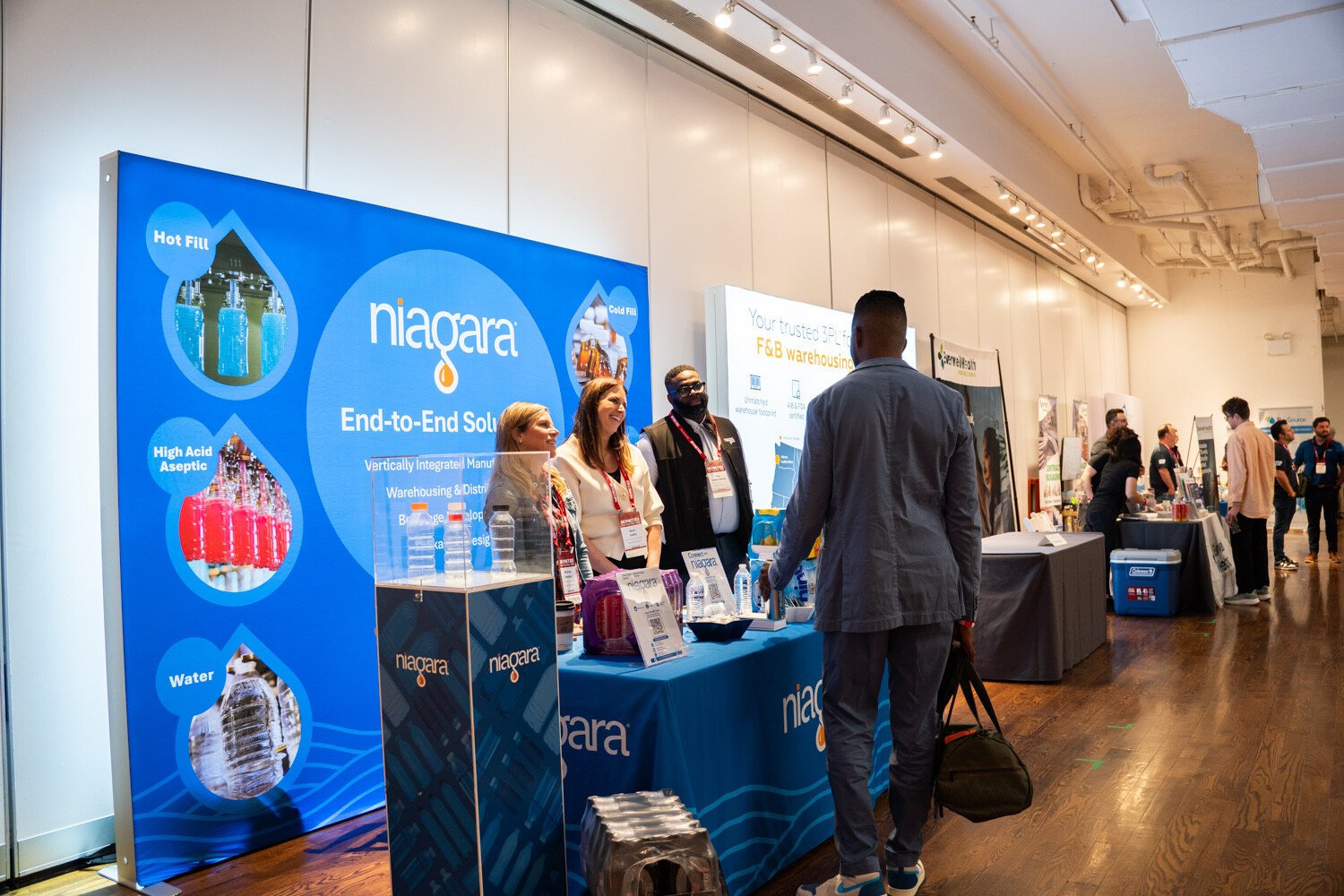 Wide view of a blue illuminated Niagara booth with staff and attendees, and more booths down the hall.