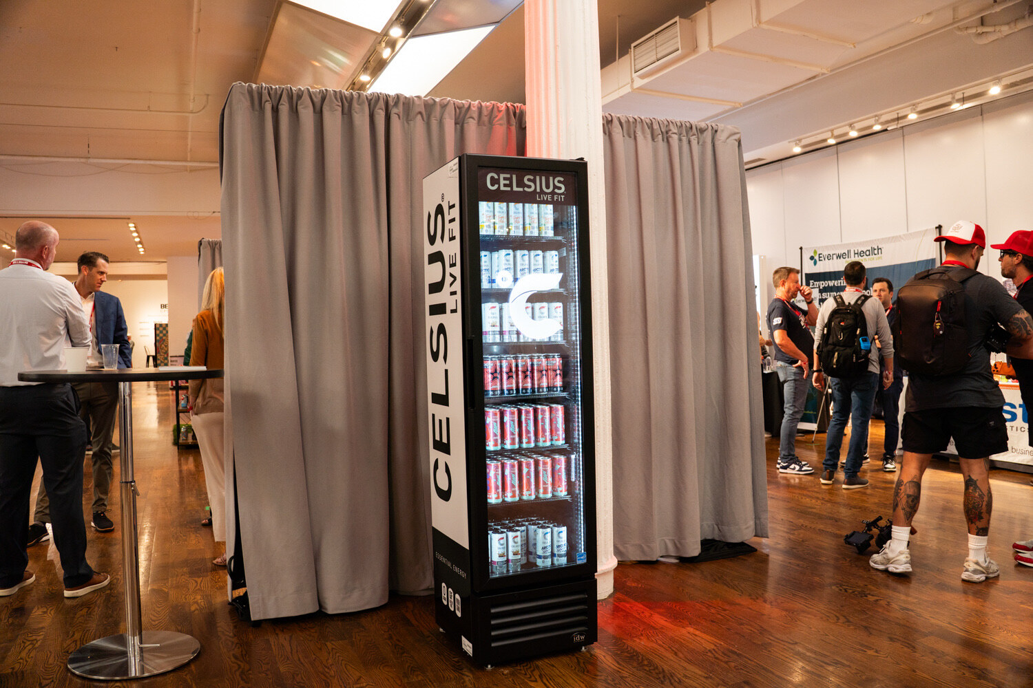A tall black Celsius fridge between gray curtains, with attendees mingling behind it.