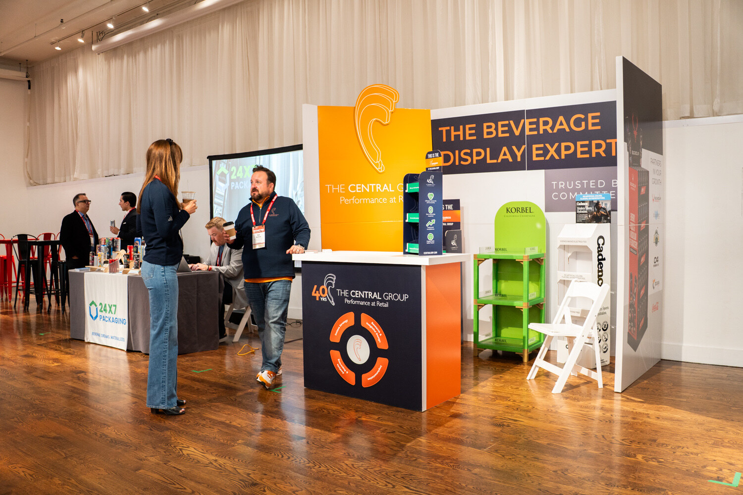 Wide view of an orange and navy display booth, two attendees, and a 24x7 Packaging table in the background.