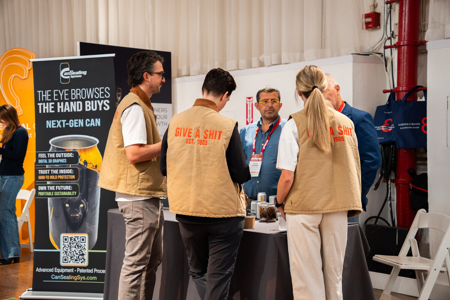 Group of attendees, two in tan vests, gather around a table beside a black banner showing a next-gen can.