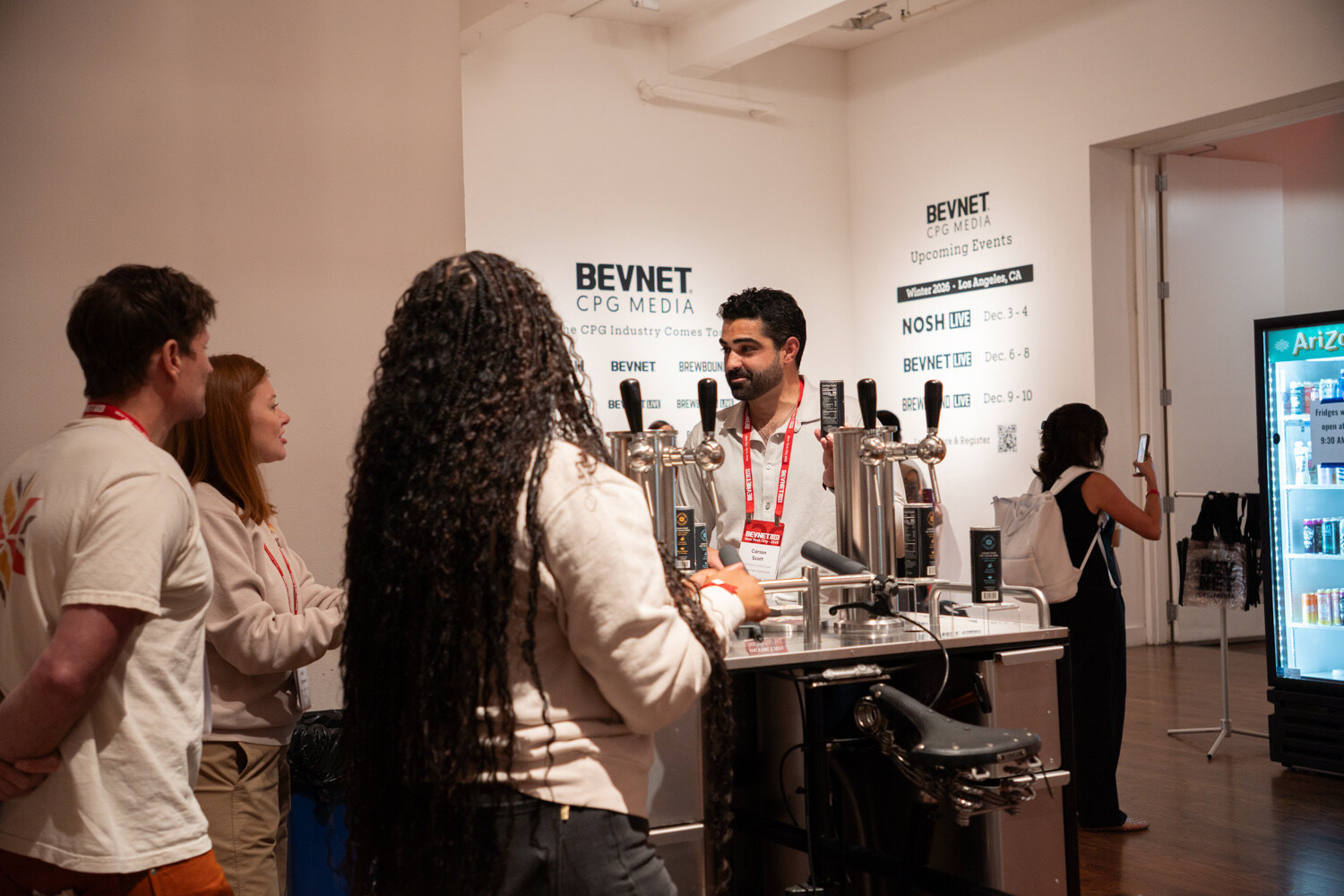 Man behind a steel tap cart serves attendees in front of a white wall of BevNET CPG Media upcoming-event graphics.