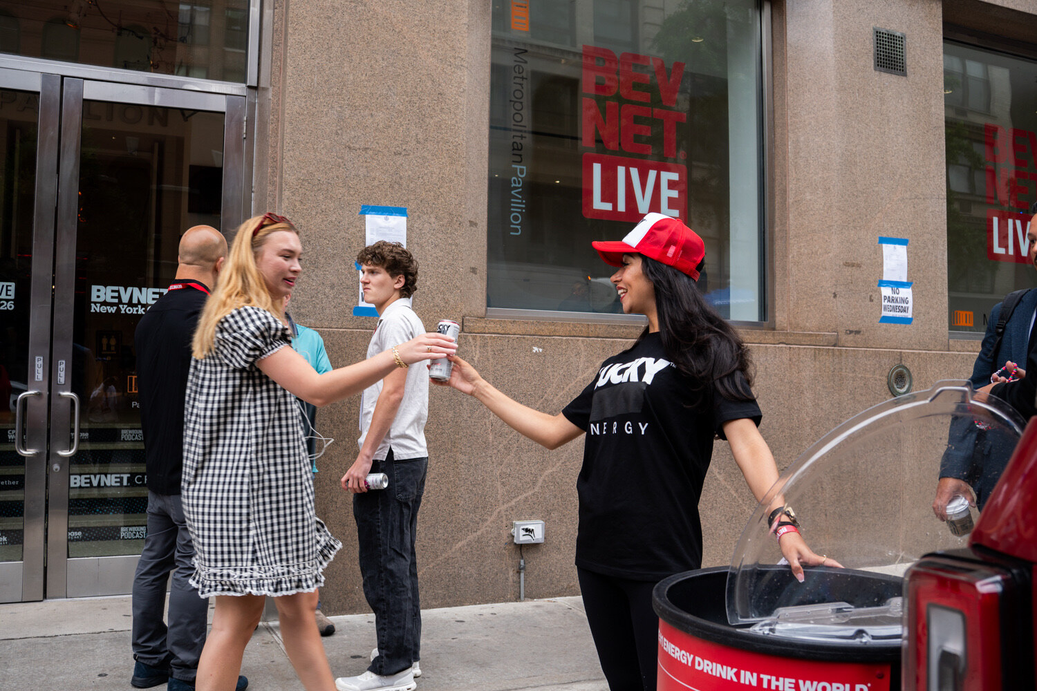 Woman in a red cap and Lucky Energy T-shirt hands a can to an attendee near a BevNET Live sign on the building.
