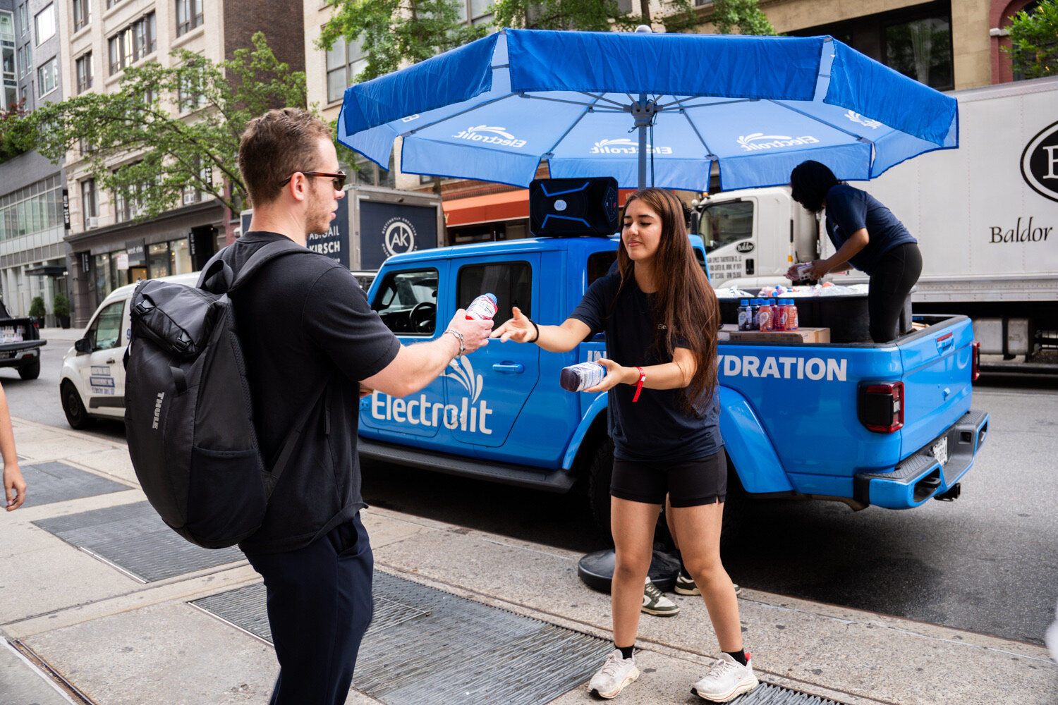 Young woman hands a bottle to a man in sunglasses beside a blue Electrolit pickup under a blue umbrella.