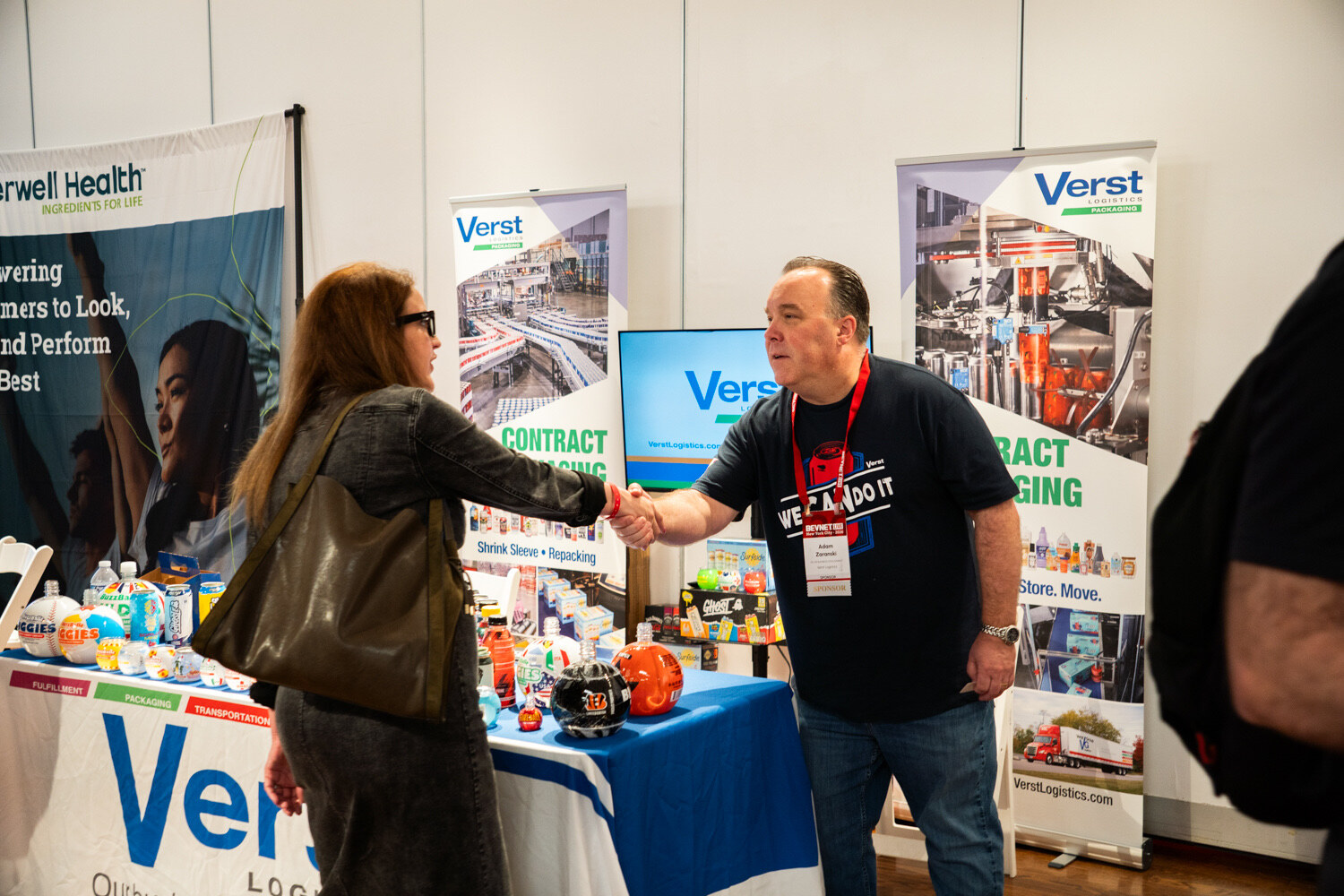Man in a navy T-shirt shakes hands with a woman across a Verst Logistics table lined with cans and bottles.