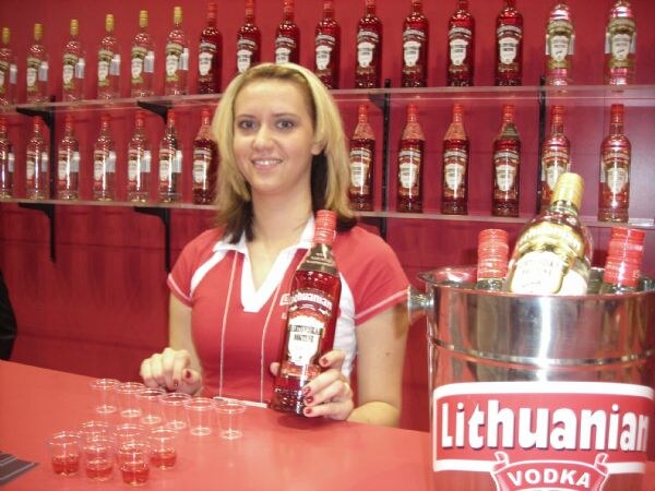 A smiling woman in a red polo holds a bottle of Lithuanian Vodka at a red counter with small sample cups, shelves of bottles behind her, and an ice bucket marked Lithuanian Vodka.