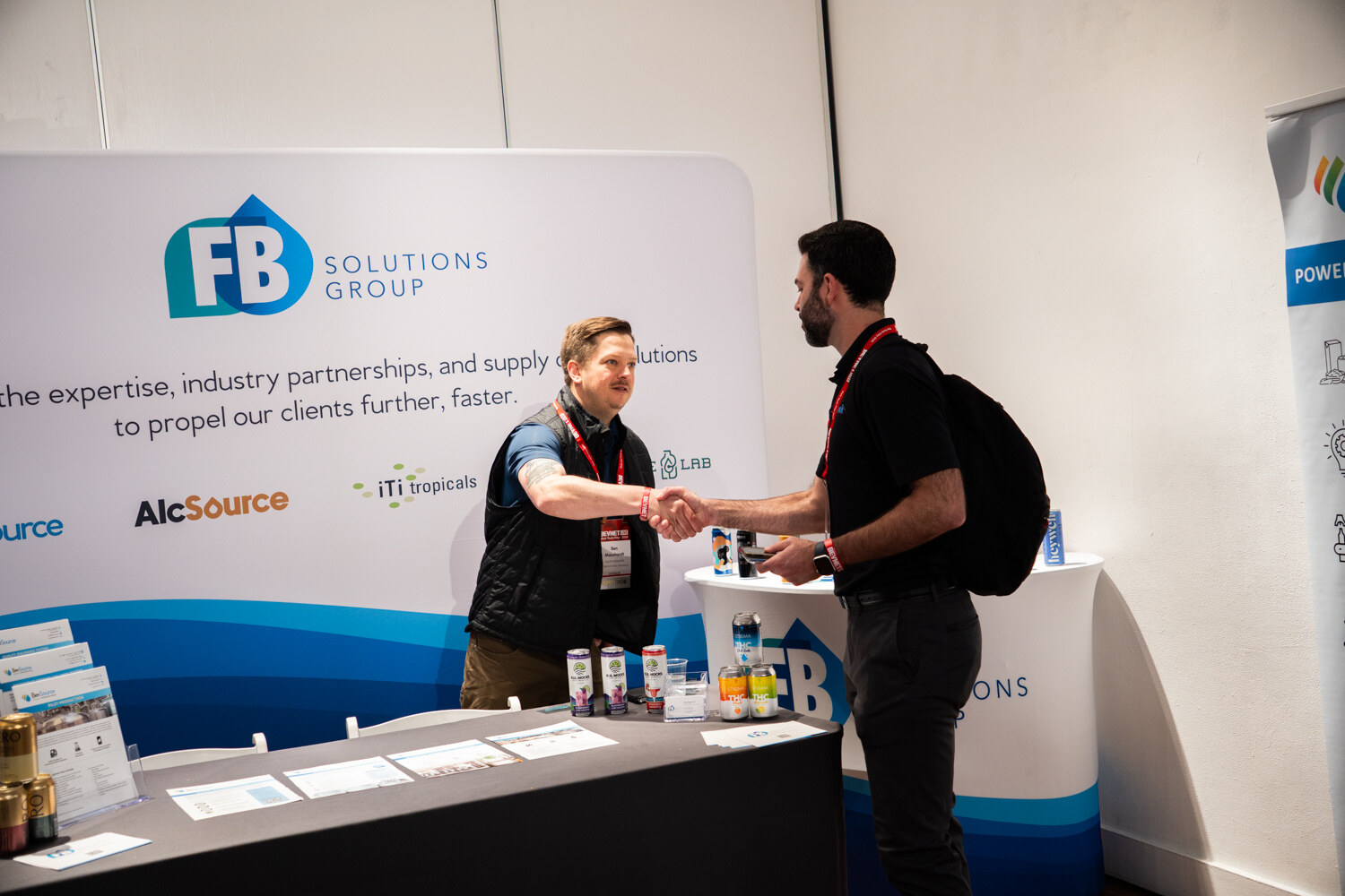 Two men shake hands over a table in front of an FB Solutions Group backdrop listing BevSource and AlcSource.
