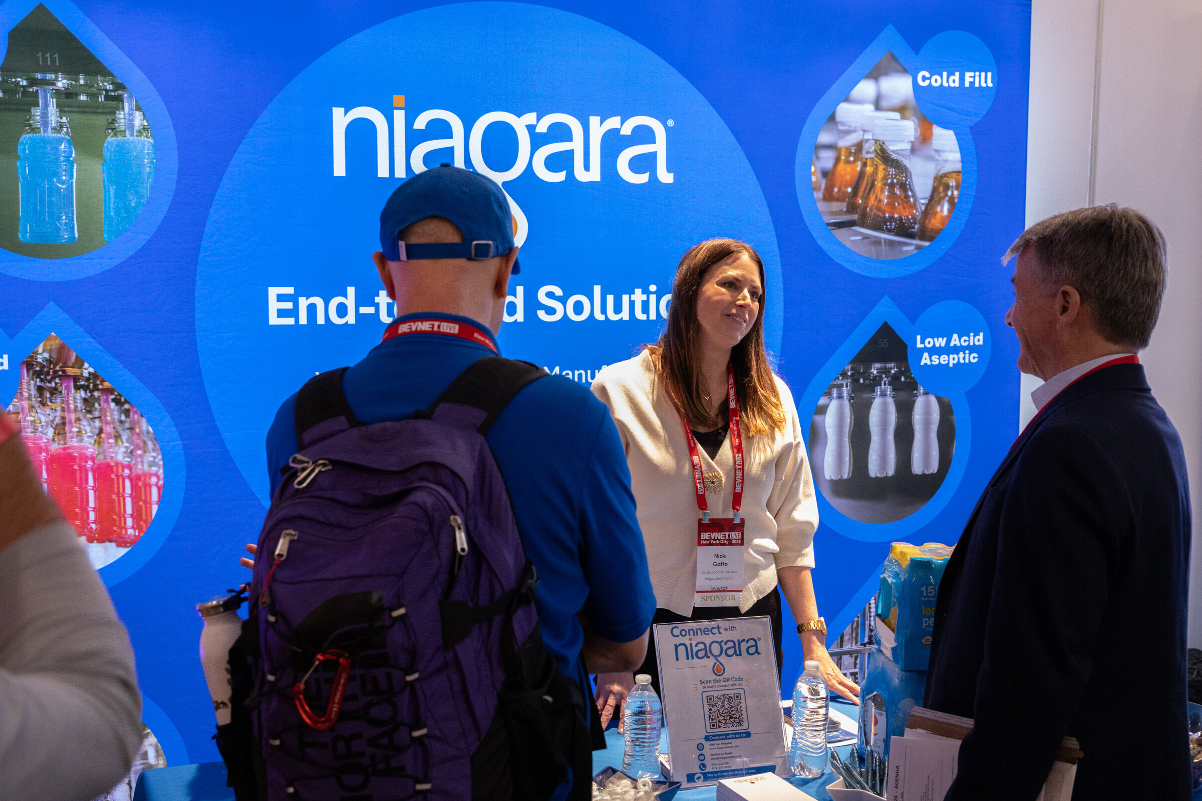 Smiling woman behind a table talks with two men in front of a bright blue Niagara backdrop.