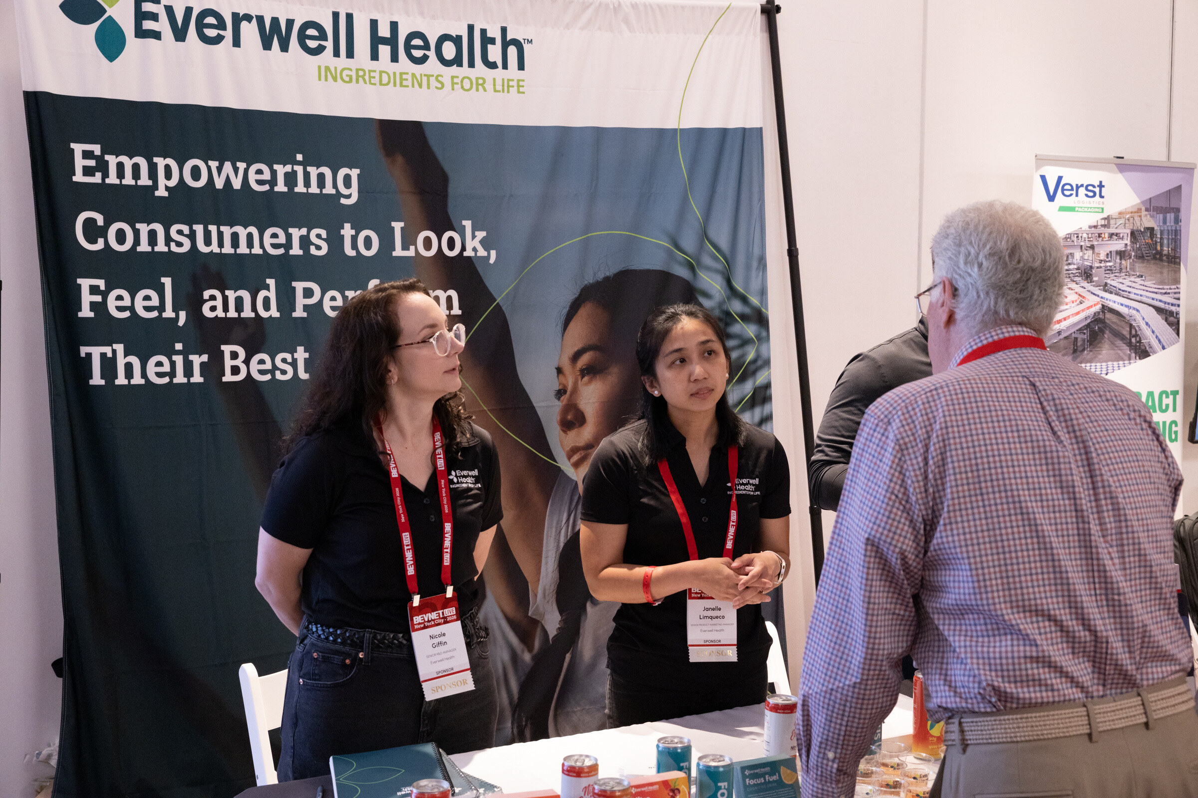 Two women in black polos listen to a gray-haired man at a table in front of an Everwell Health banner.