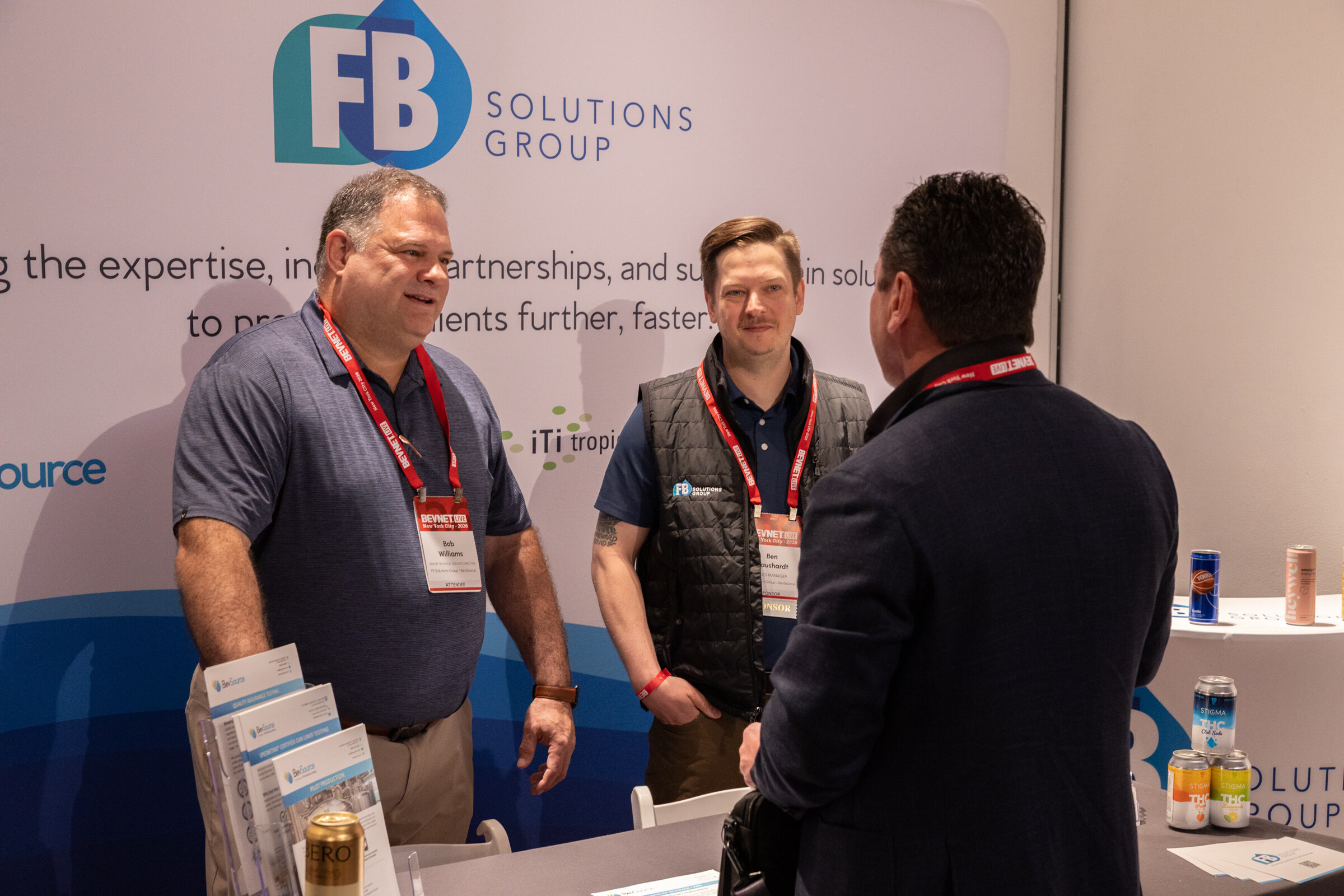 Two smiling men in lanyards talk with an attendee in front of a white FB Solutions Group backdrop.