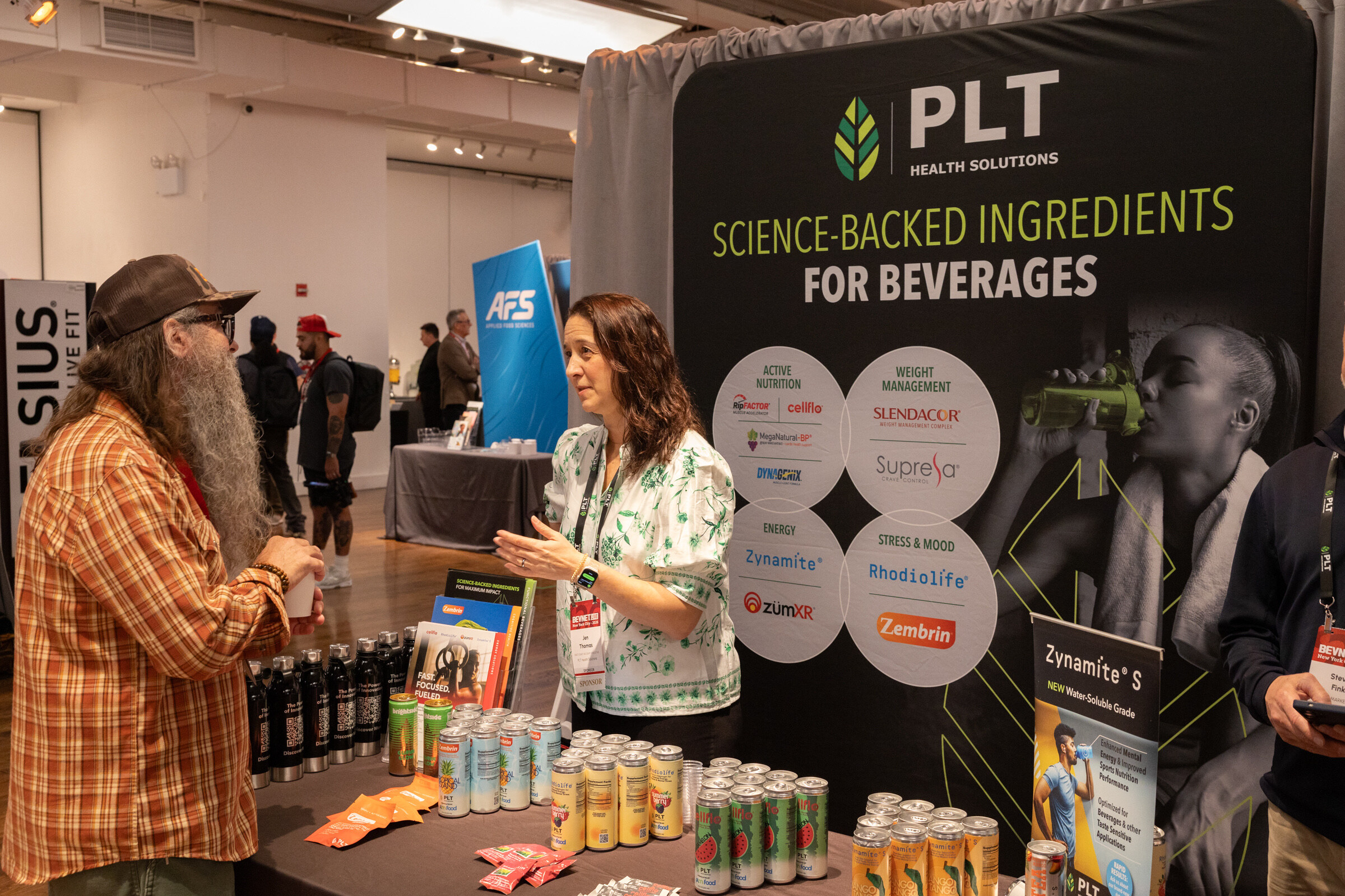 Woman in a floral blouse gestures while talking with a bearded man in a plaid shirt across a table of cans, in front of a black PLT backdrop.