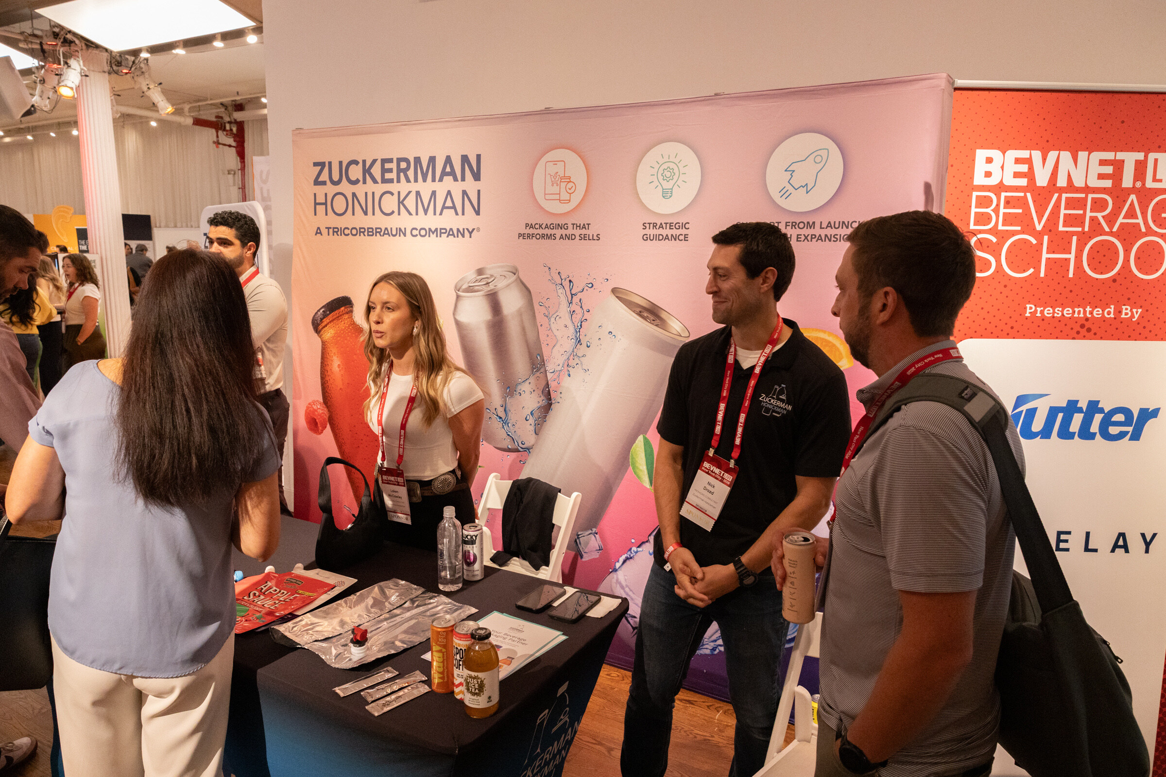 Two Zuckerman Honickman staff talk with attendees at a table of bottles, in front of a pink backdrop of cans and bottles.
