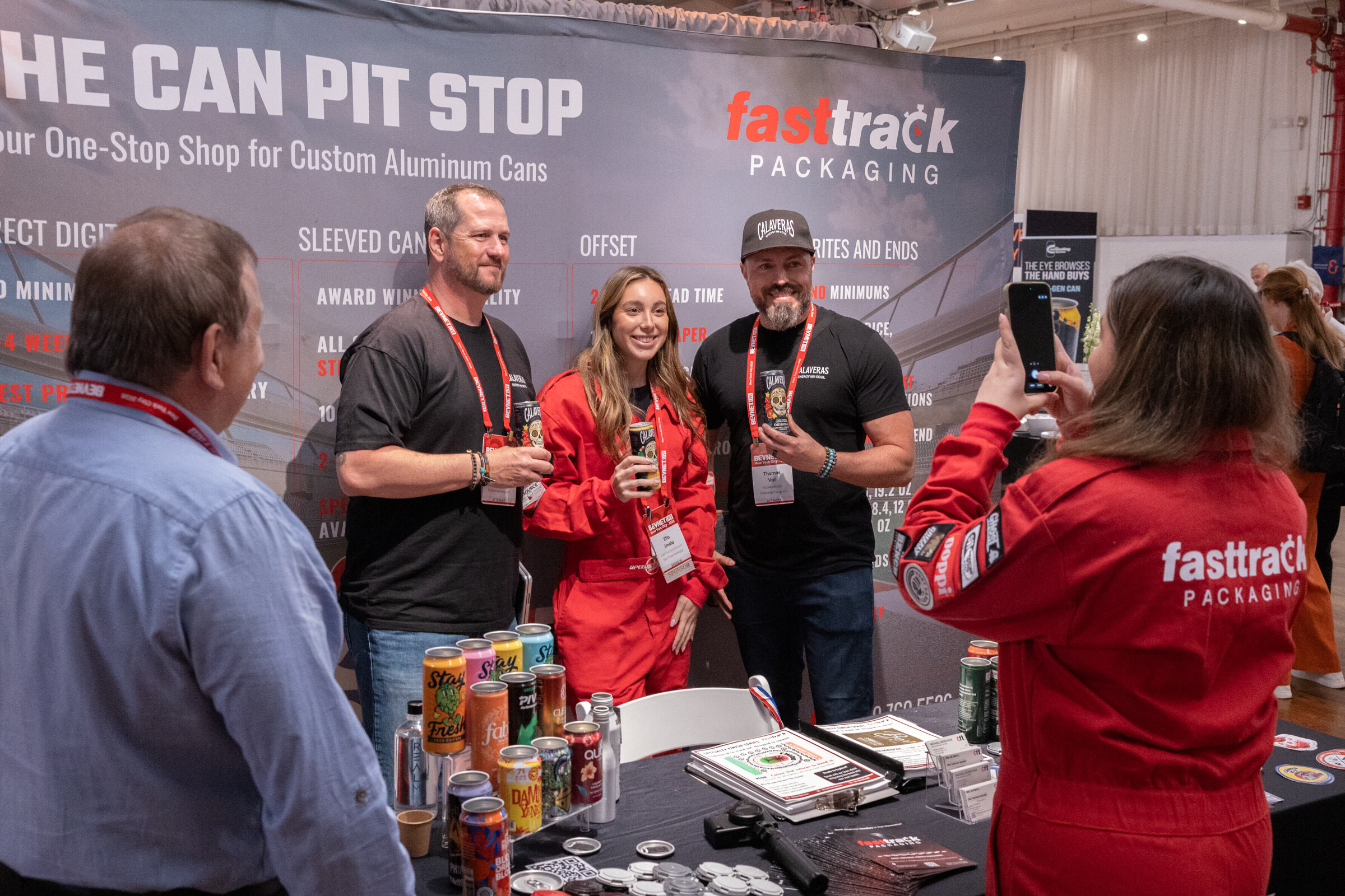 Two men and a woman in a red jumpsuit hold cans in front of a Can Pit Stop backdrop while a woman in a red Fasttrack Packaging jumpsuit photographs them.