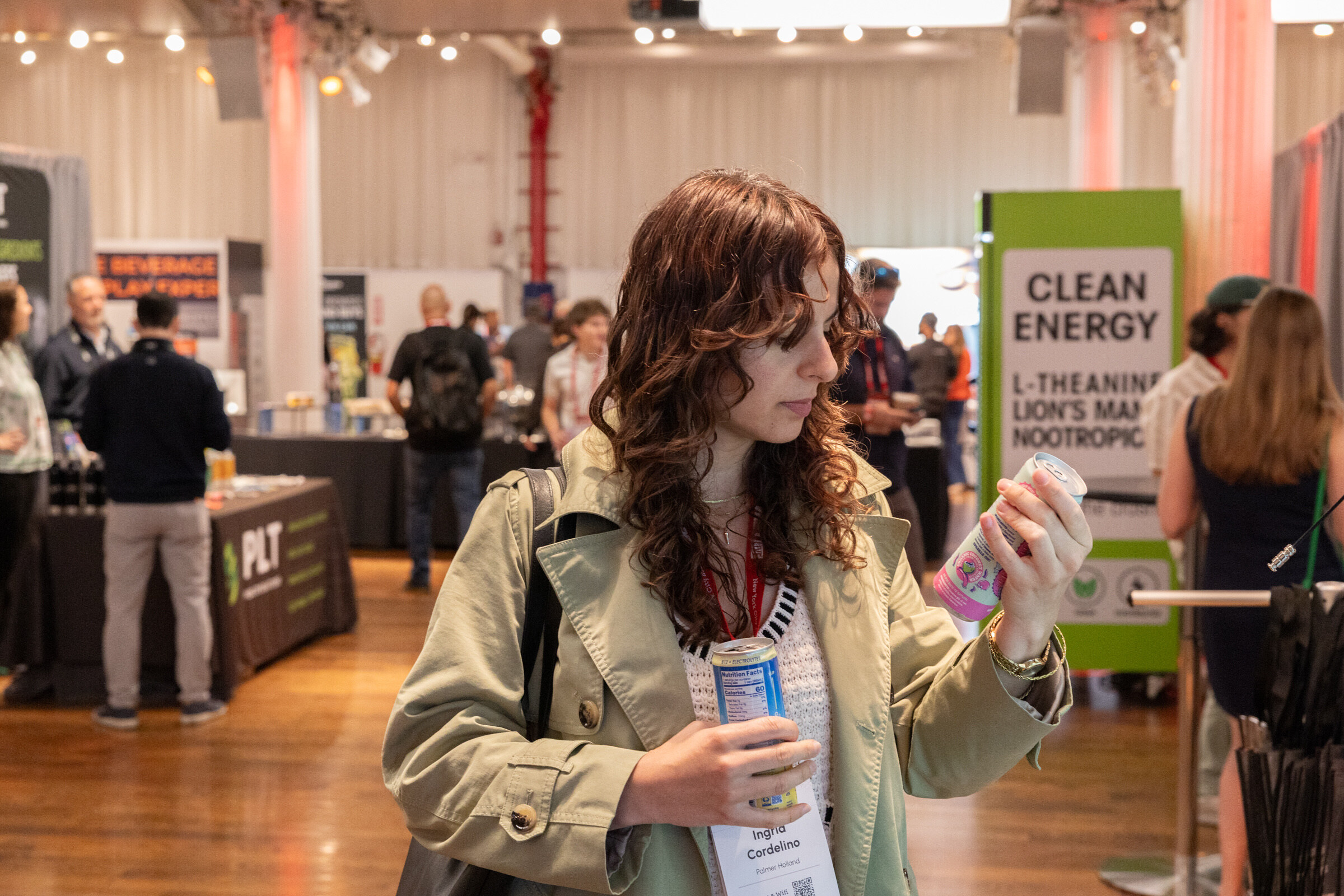 Woman with curly hair in a green trench coat reads a pink can while holding another, with booths behind her.