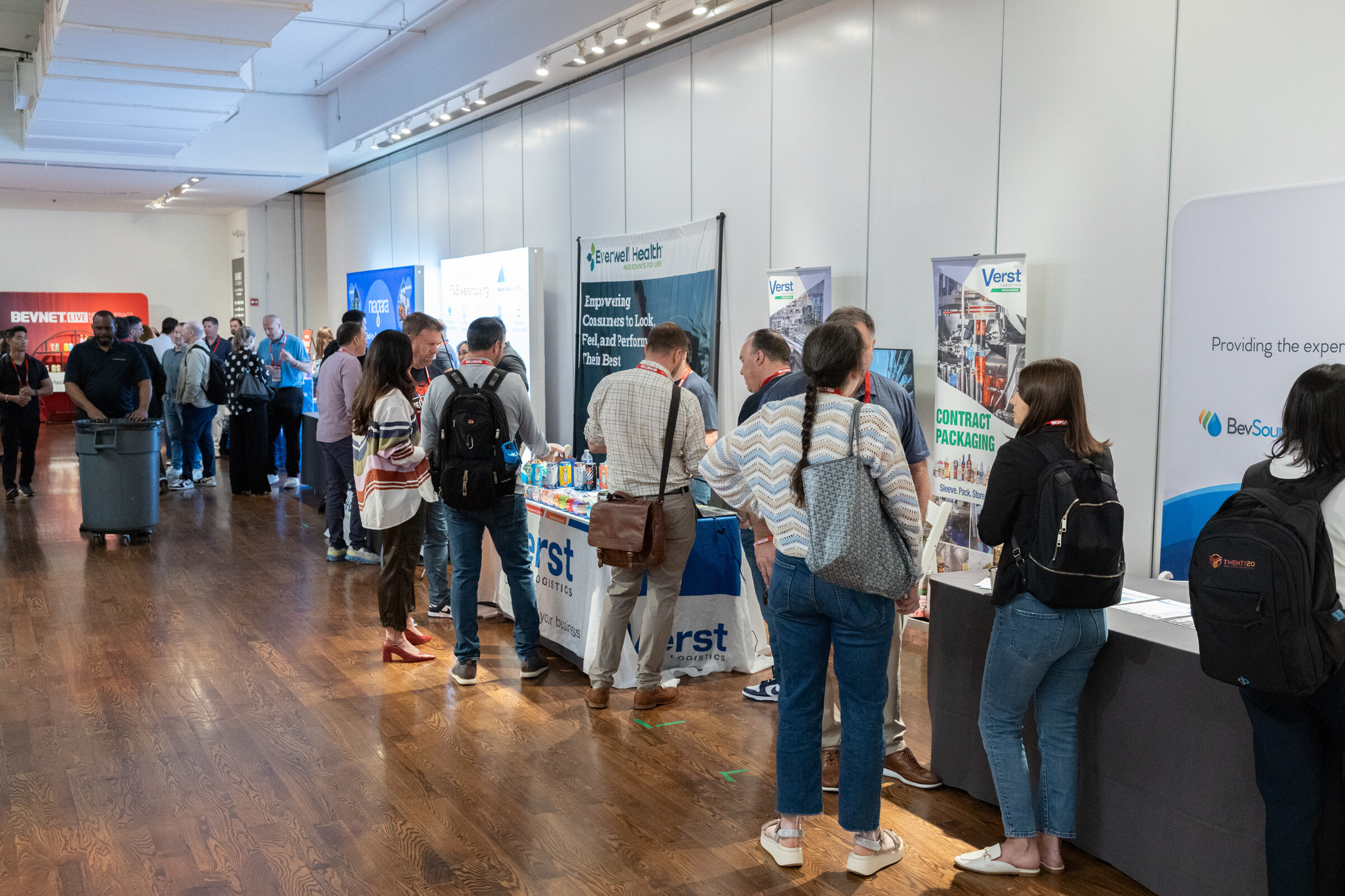 Attendees walk and talk along a row of sponsor booths in a bright hall with wood floors.