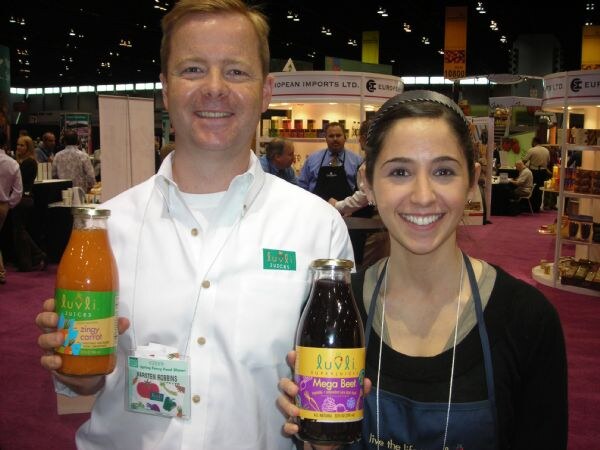 A man in a white shirt holding a Luvli Zingy Carrot juice and a woman in an apron holding a Luvli Mega Beet juice smile in front of other booths on a trade show floor.